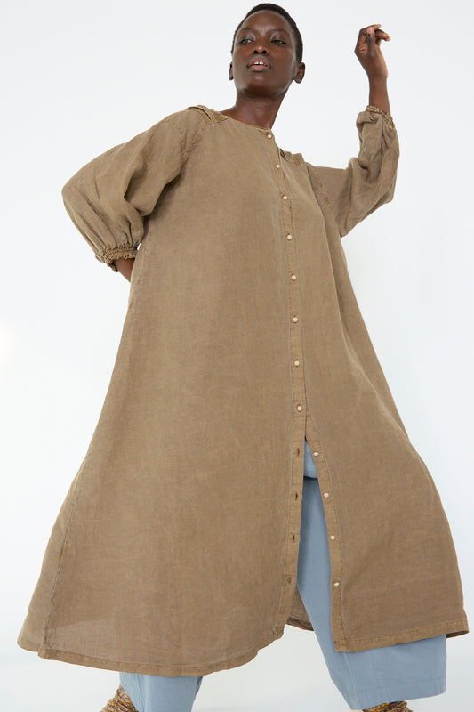 A person is wearing the Natural Dyed Linen Cotton Lace Dress in Camel by nest Robe over light blue pants, and is standing with one hand raised against a white background.