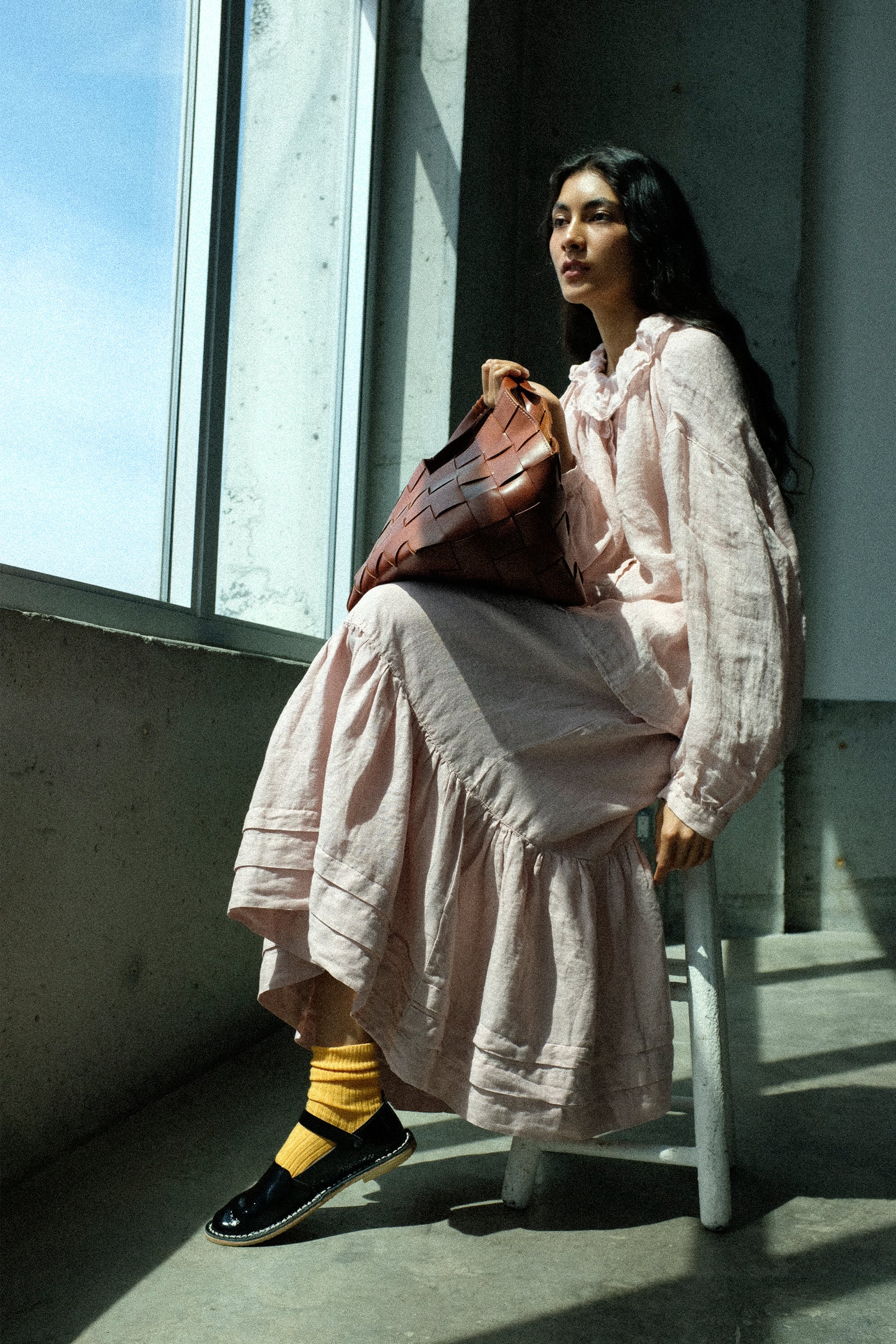 Woman in wearing nest Robe's Linen Gauze Frill Blouse in Light Peach, and Linen Natural Dyed Pleated Gathered Skirt in Pink sitting by a window with sunlight filtering through, holding the Dragon Diffusion Botteghe Bag in Tan.