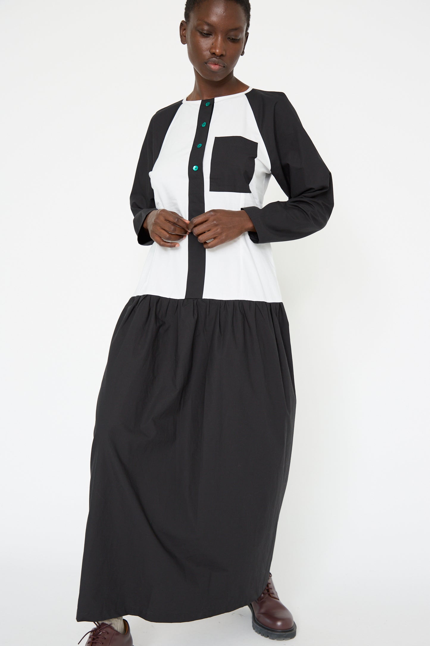 A woman is standing against a plain white background wearing the Cotton Poplin Frame Dress in Flash by SC103, a relaxed-fit maxi dress featuring black and white colors with green buttons.