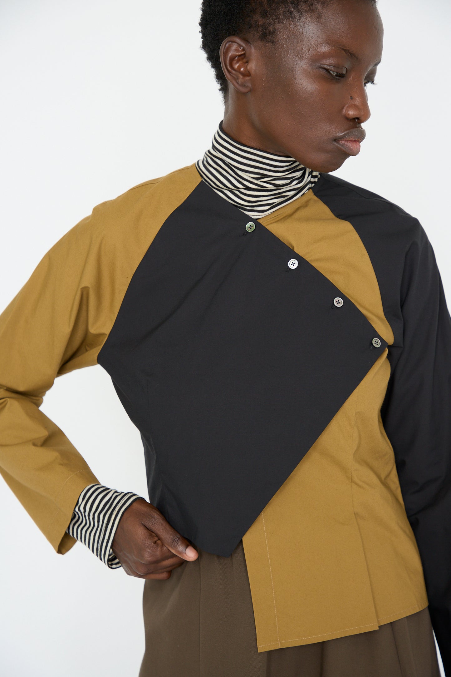 A woman in the Cotton Poplin Peak Blouse by SC103, showcasing a geometric pattern and an asymmetrical hem, pairs it with a striped turtleneck underneath while looking down to the side.