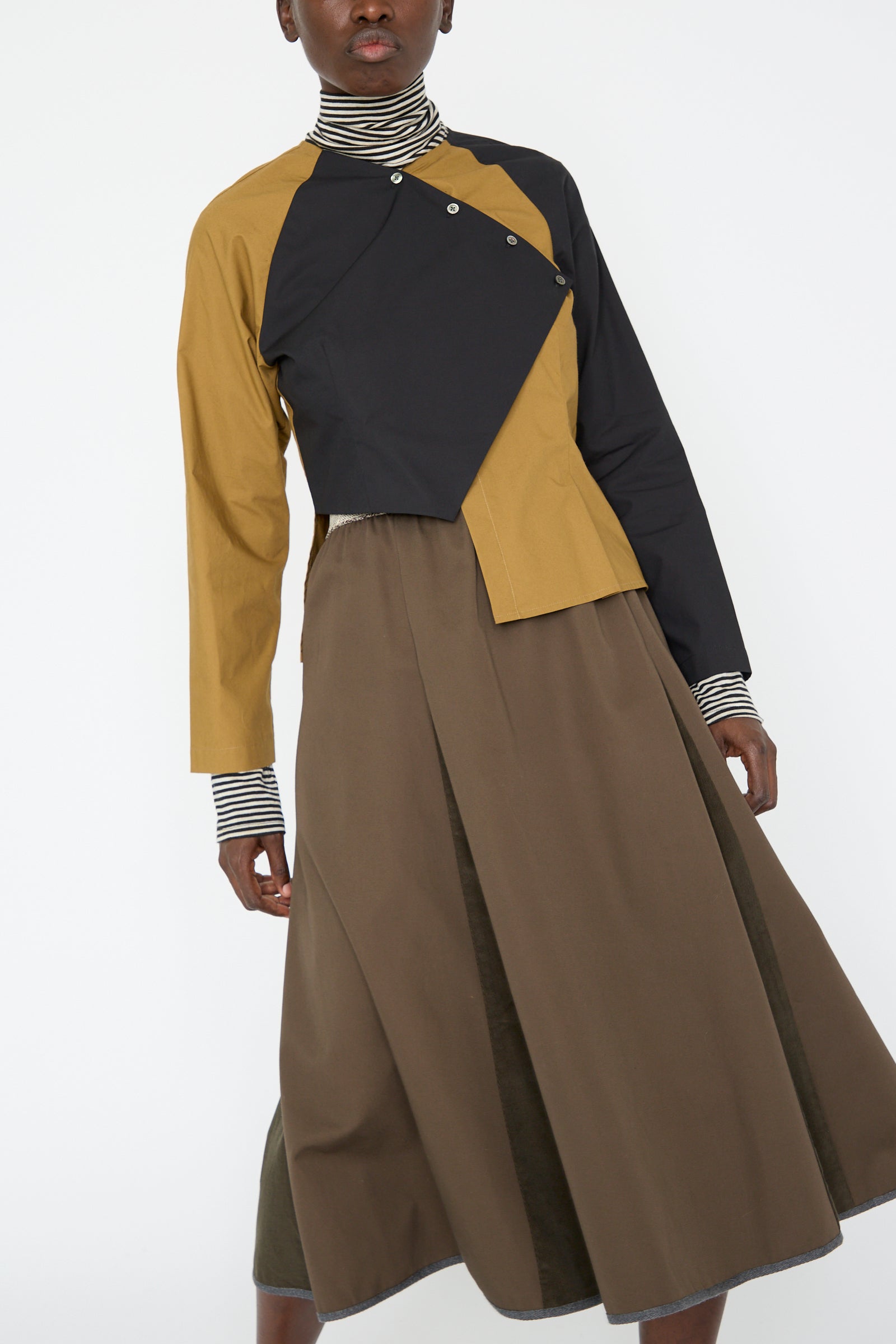 A woman wearing the SC103 Cotton Poplin Peak Blouse in Oblique, featuring color-blocked sections of black and mustard and an asymmetrical hem, stands against a plain background, with a brown skirt.