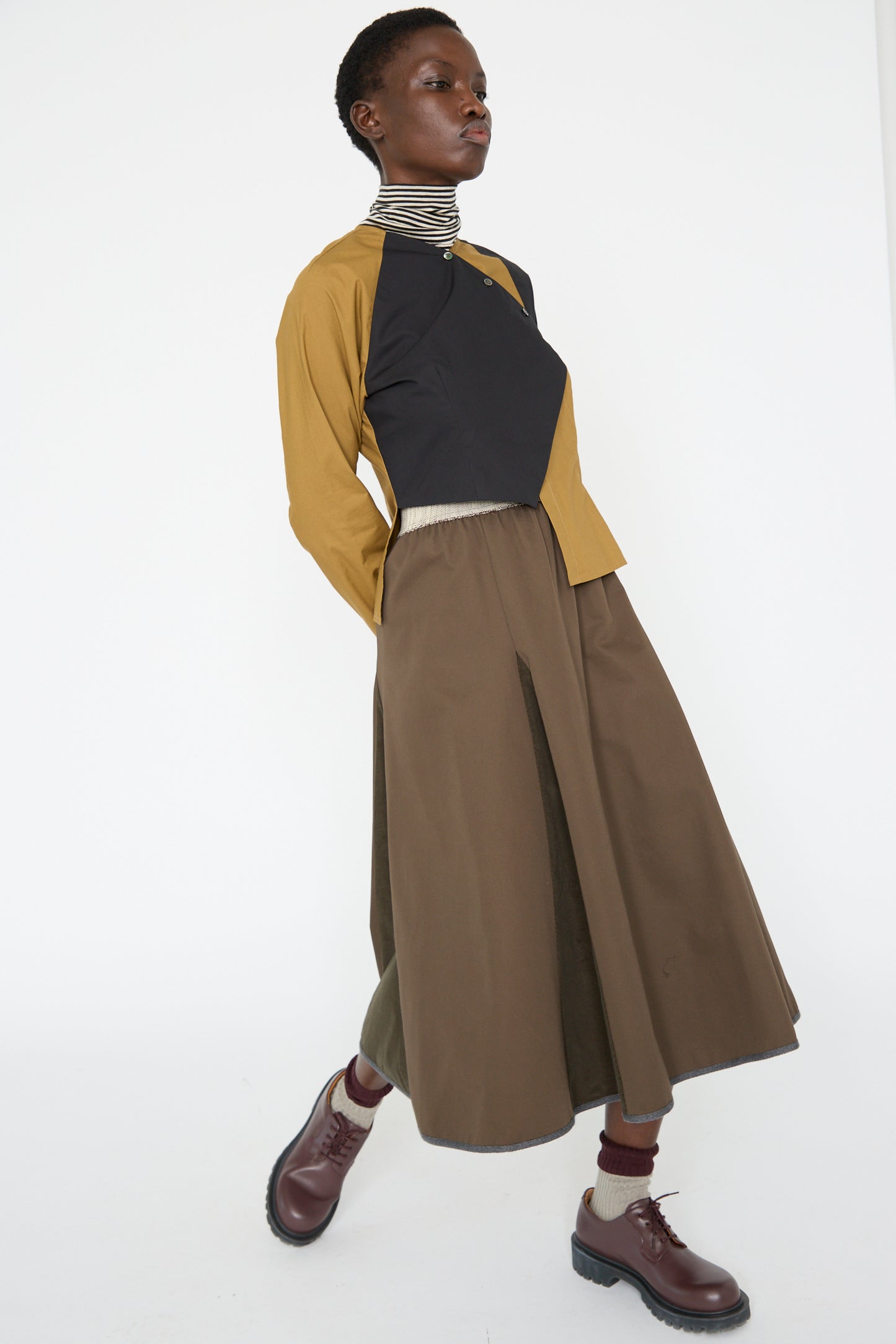 A woman walks to the left wearing the SC103's Cotton Twill Axis Skirt in Beam, paired with a two-tone top and brown shoes on a plain white background.