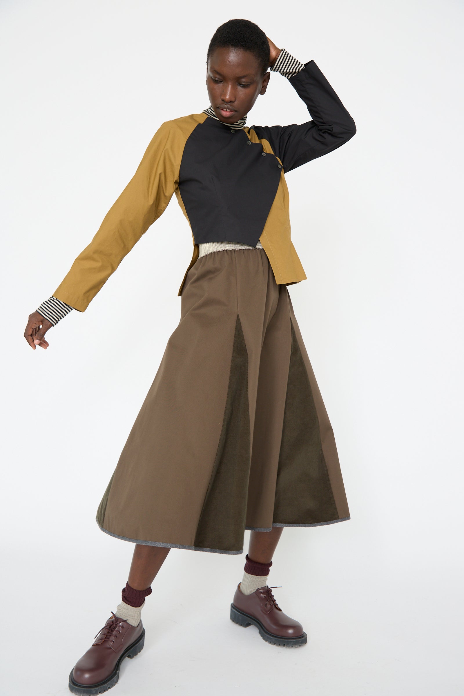 A woman wearing a two-toned top paired with SC103's Cotton Twill Axis Skirt in Beam. She strikes a pose with one arm bent above their head.