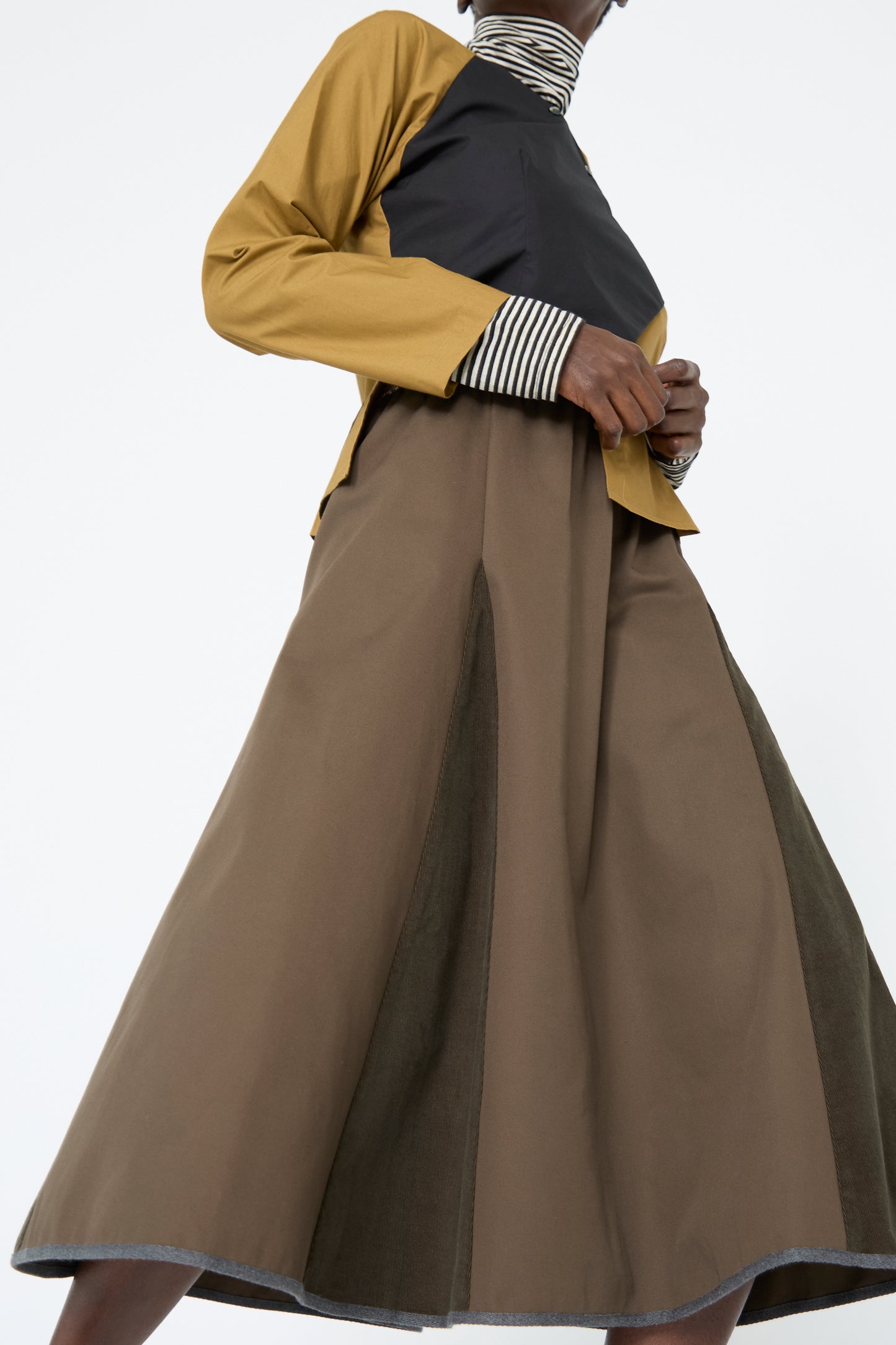 A woman wearing a yellow jacket and striped top stands against a white background, showcasing the effortlessly chic SC103's Cotton Twill Axis Skirt in Beam. 