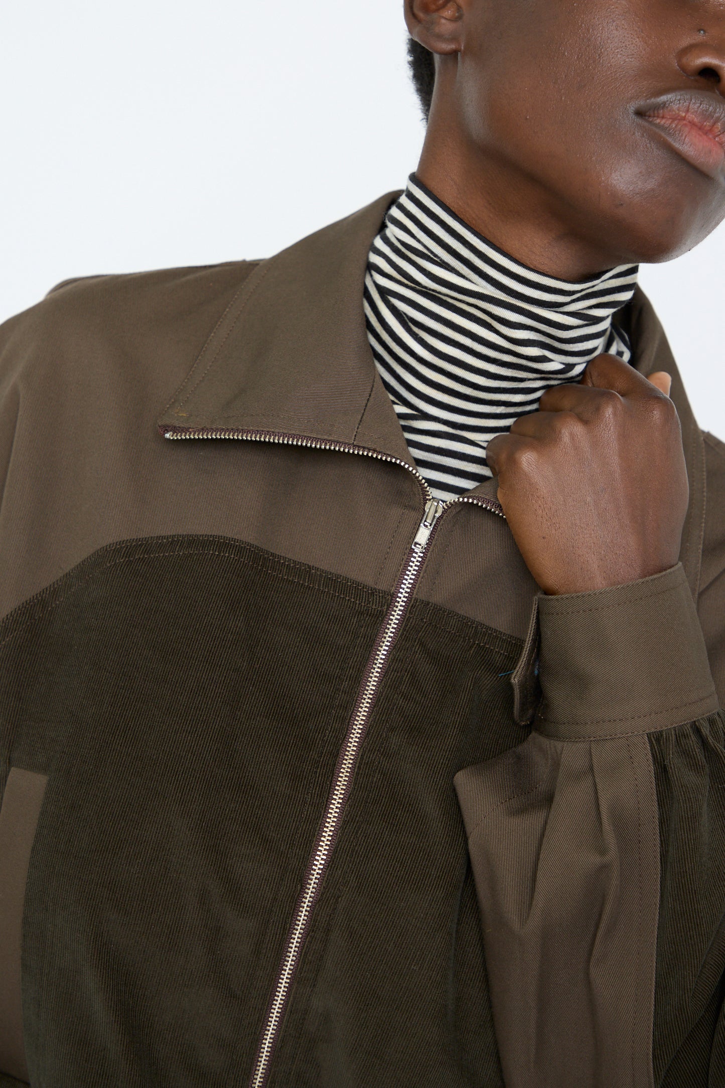 A woman wearing the SC103 Cotton Twill Coda Jacket in Beam, a relaxed-fit brown jacket with a zipper, over a black and white striped turtleneck.