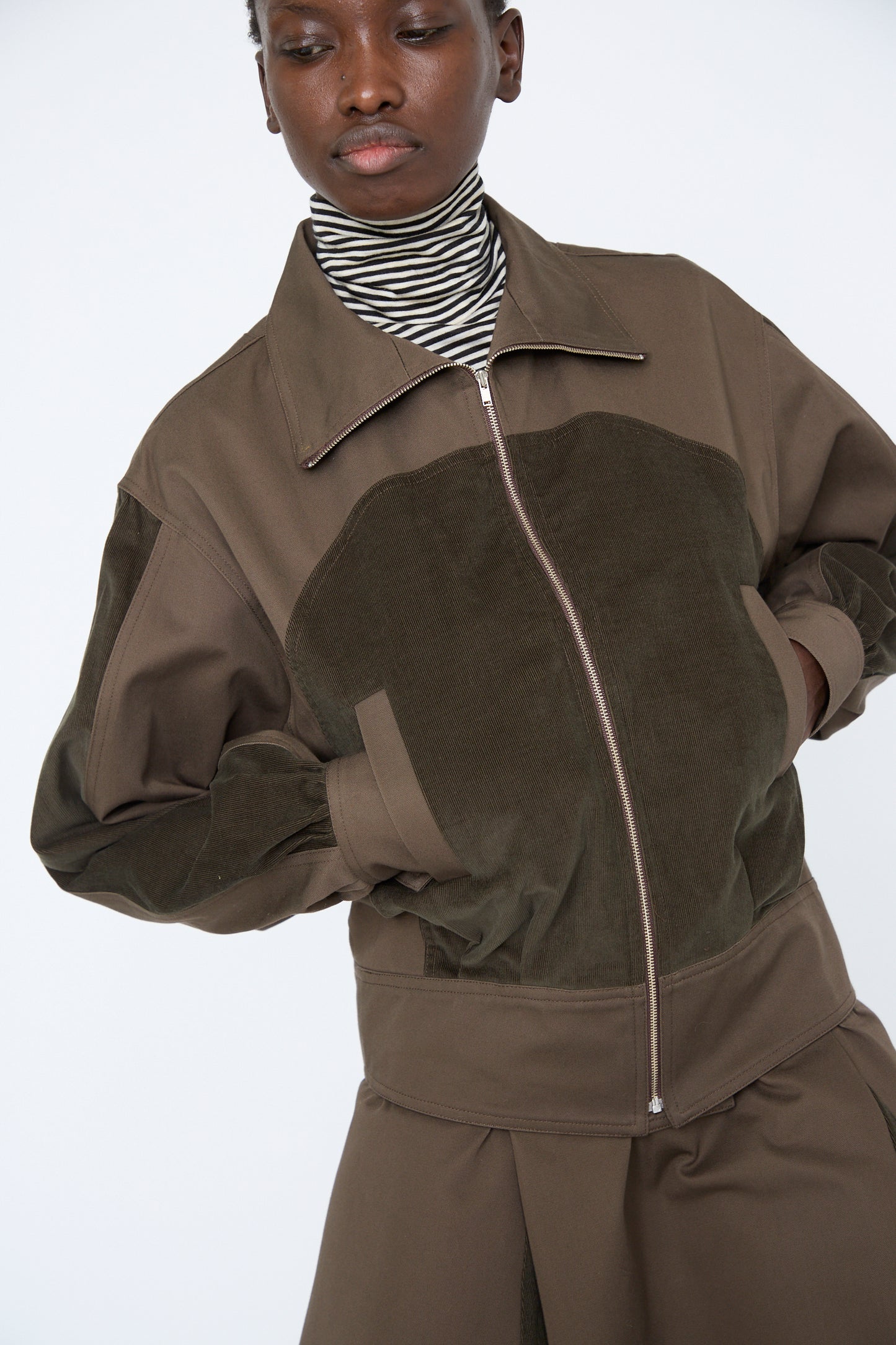 A woman wearing the Cotton Twill Coda Jacket in Beam by SC103, a relaxed fit olive green zip-up jacket featuring a high collar, paired with a striped turtleneck, looking down.