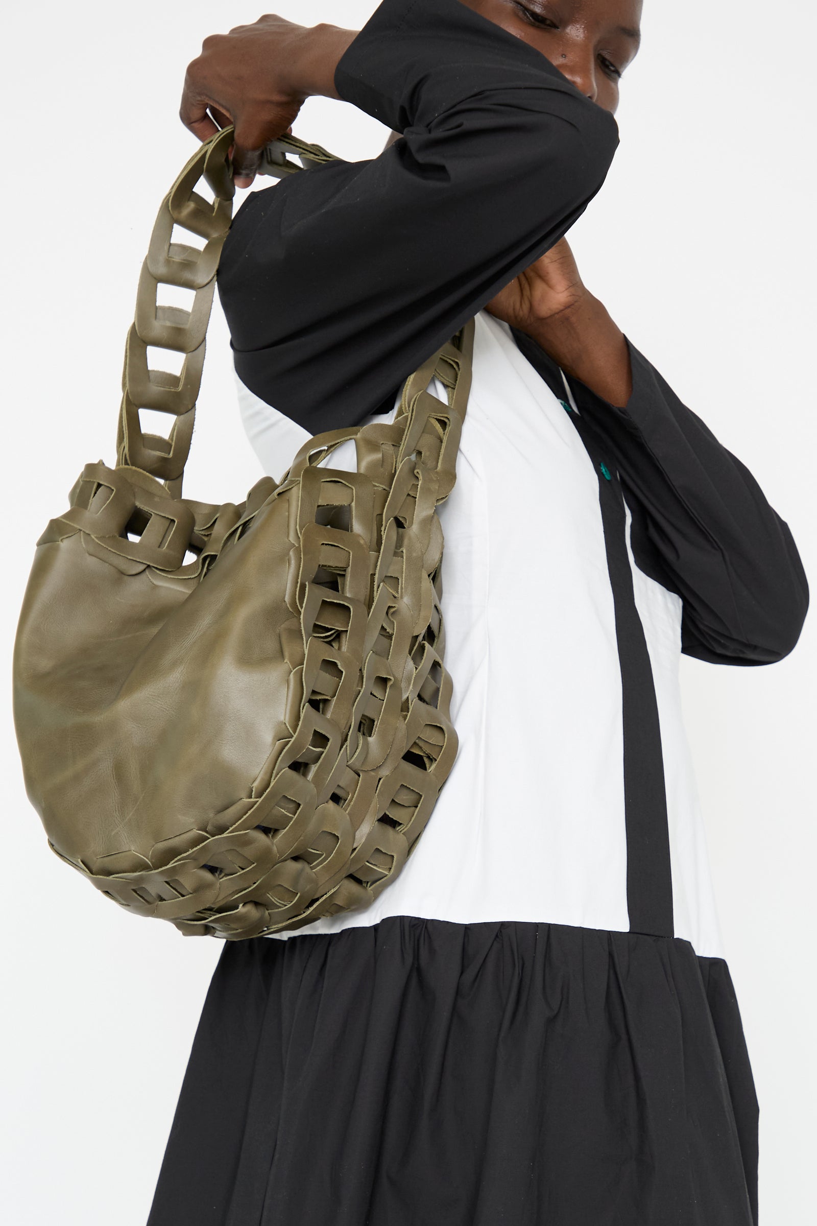 A woman dressed in black and white is carrying the SC103 Leather Drum Bag in Olive, characterized by its handmade design and chain-link strap.