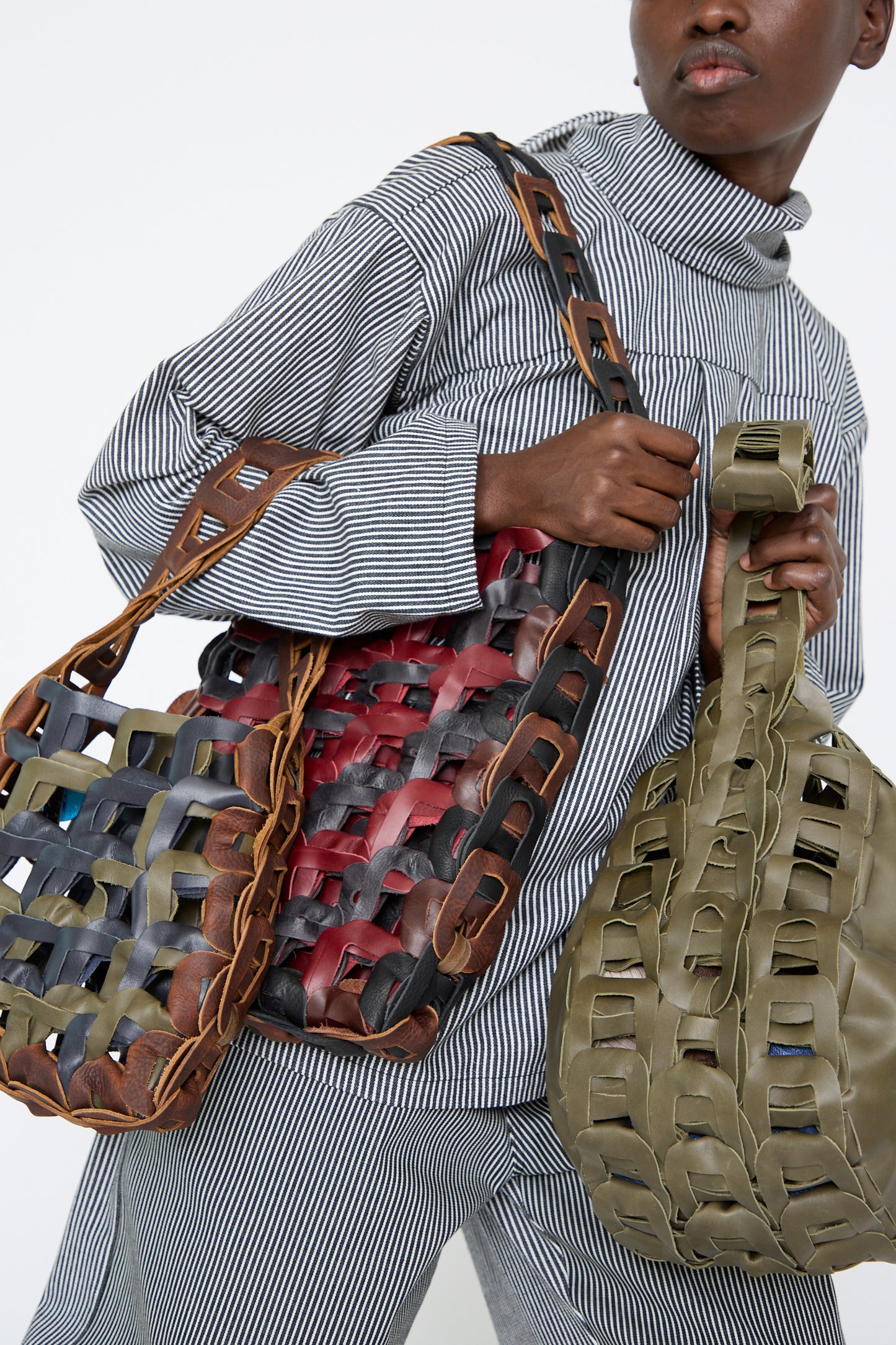 A woman holding two SC103 handmade leather bags—one, the Leather Links Tote in Anise with Green, and the other in multicolored hues—styled with a black and white striped outfit.