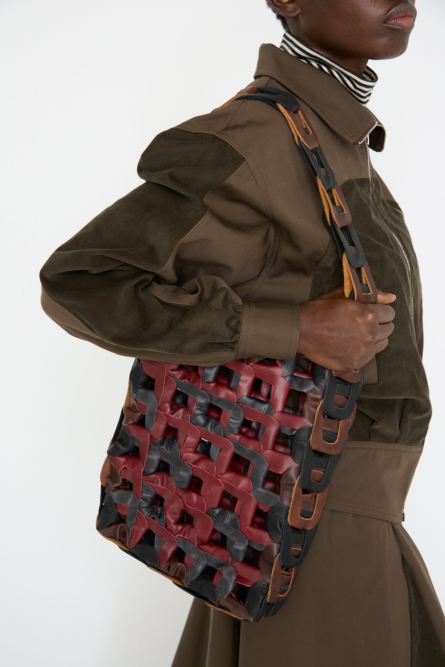 A person is holding the SC103 Leather Medium Links Tote in a Cinema design, and is dressed in a brown long-sleeve outfit with a striped collar, against a white background.