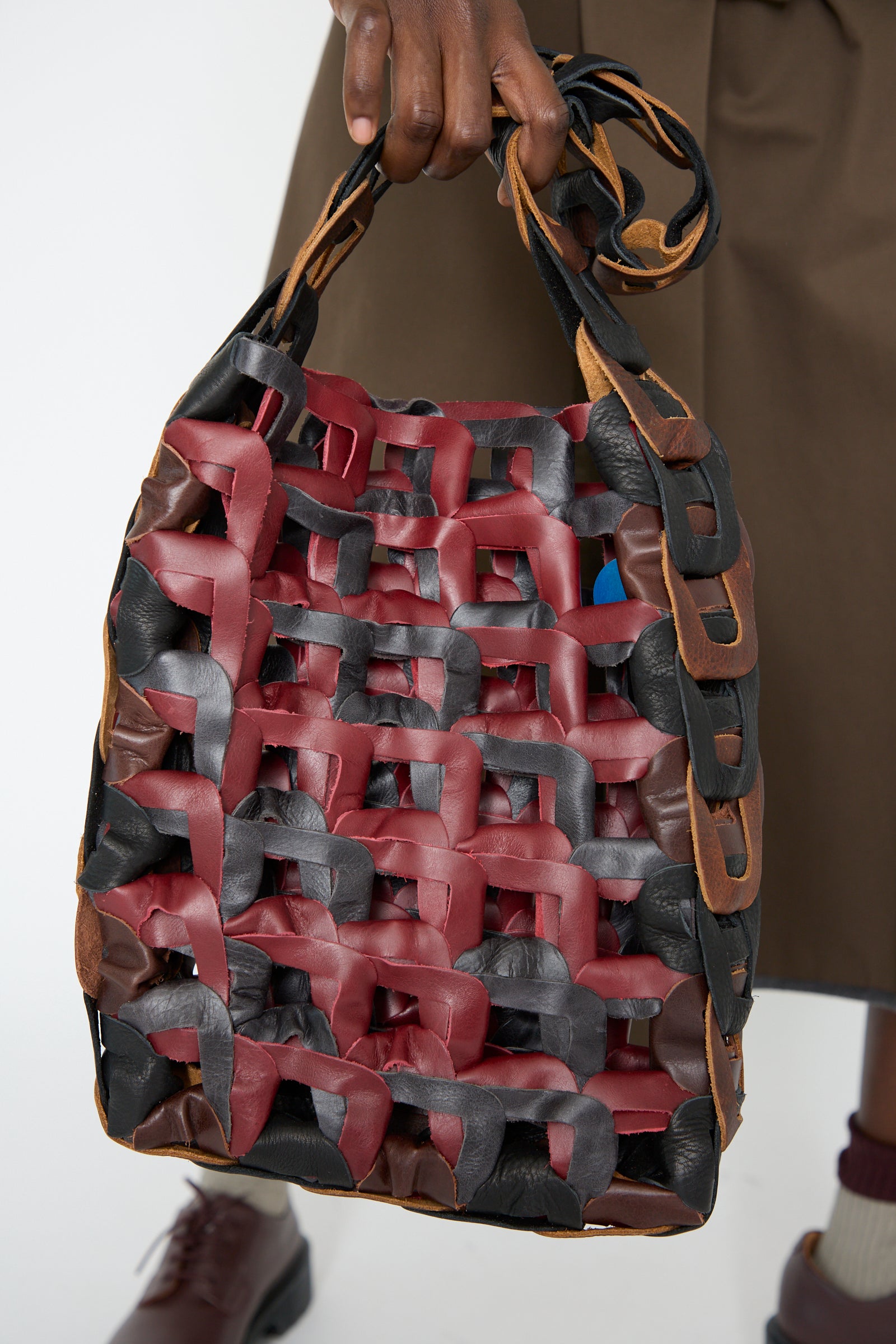 A person holds the SC103 Leather Medium Links Tote in Cinema, crafted from interlaced red, black, and brown strips, featuring a braided handle.