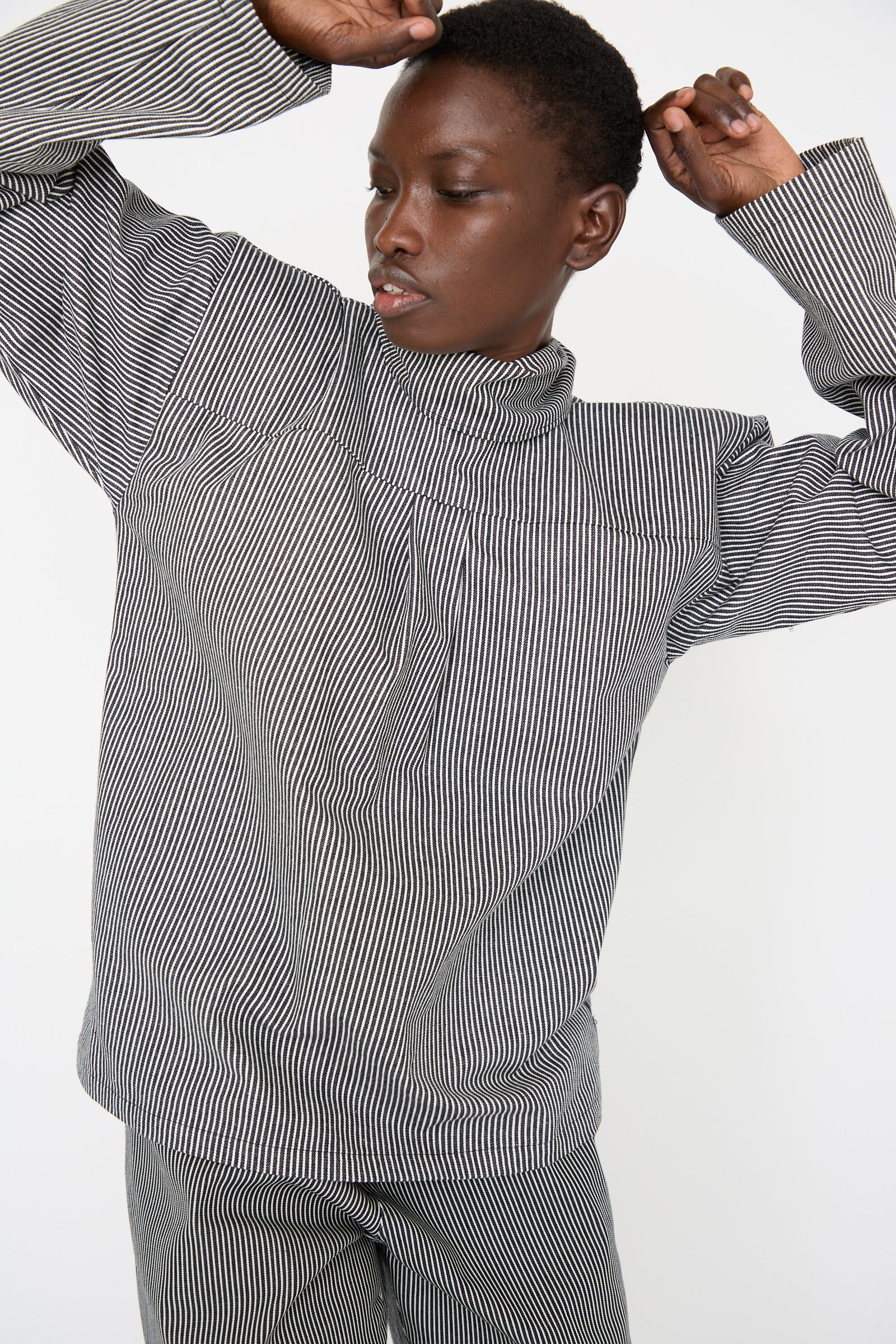 A woman models with arms raised against a white backdrop, wearing the Stripe Denim Compass Blouse in Railroad by SC103.