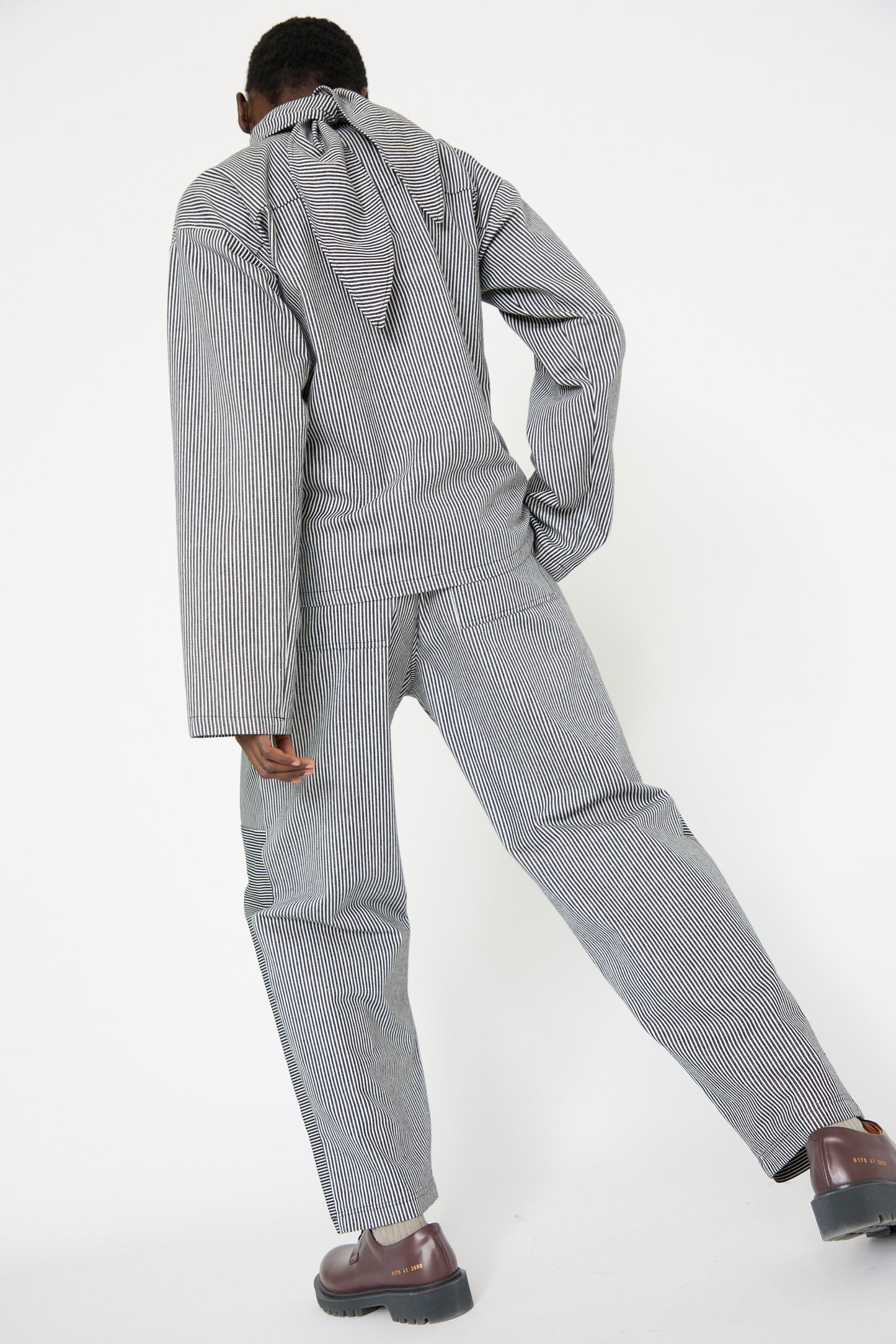 A woman is dressed in the SC103 Stripe Denim Dispatch Pant in Railroad with an elasticated waist, leaning slightly forward against a plain white background, one arm bent back and the other by their side.