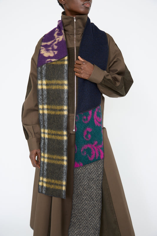 A woman wears a long brown coat, accented by the SC103 Wool Mix Ladder Scarf in Attic, featuring vibrant pattern blocks in purple, yellow, green, and pink designs.