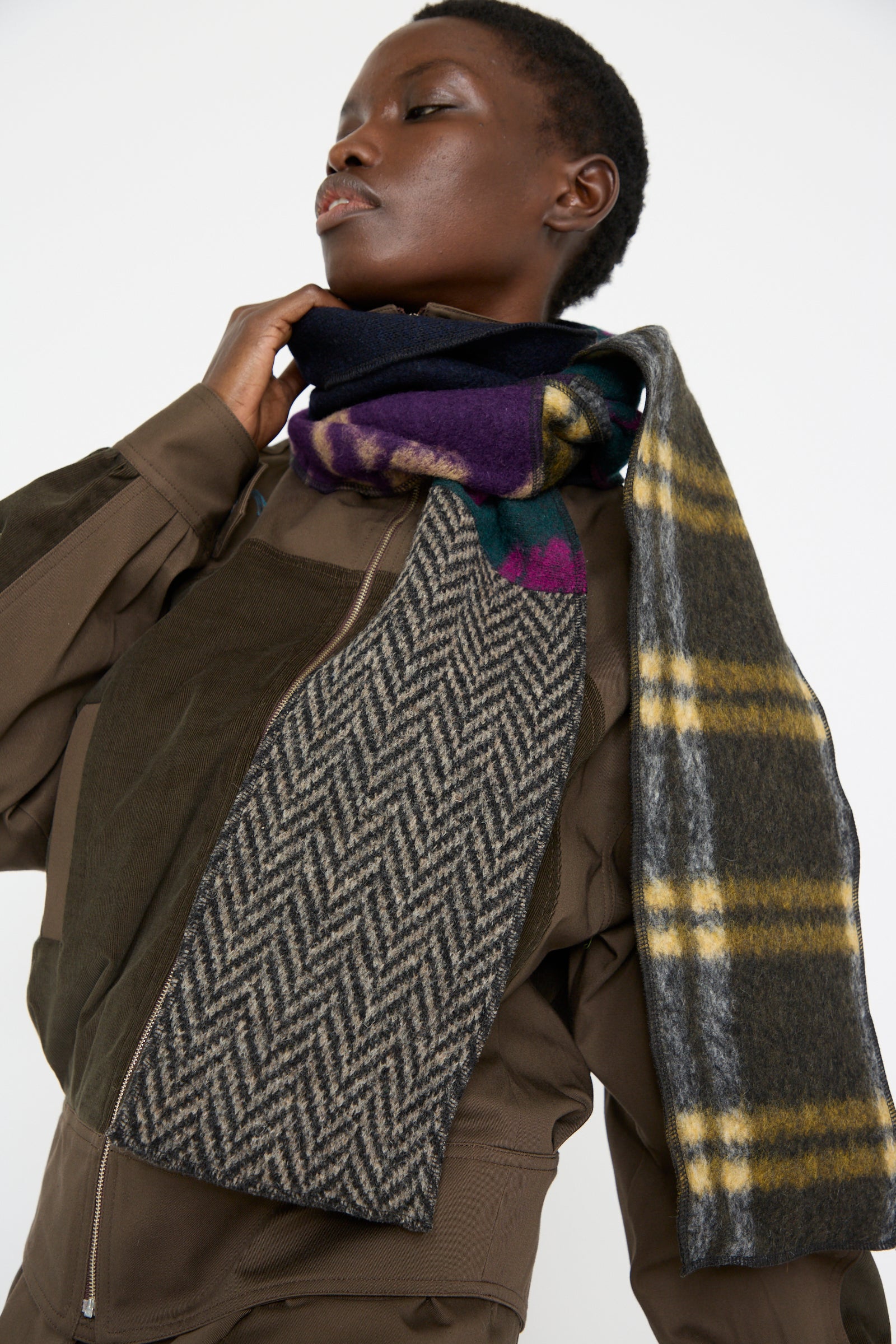 A woman wears a brown jacket complemented by the SC103 Wool Mix Ladder Scarf in Attic, showcasing plaid and herringbone pattern blocks against a plain background.