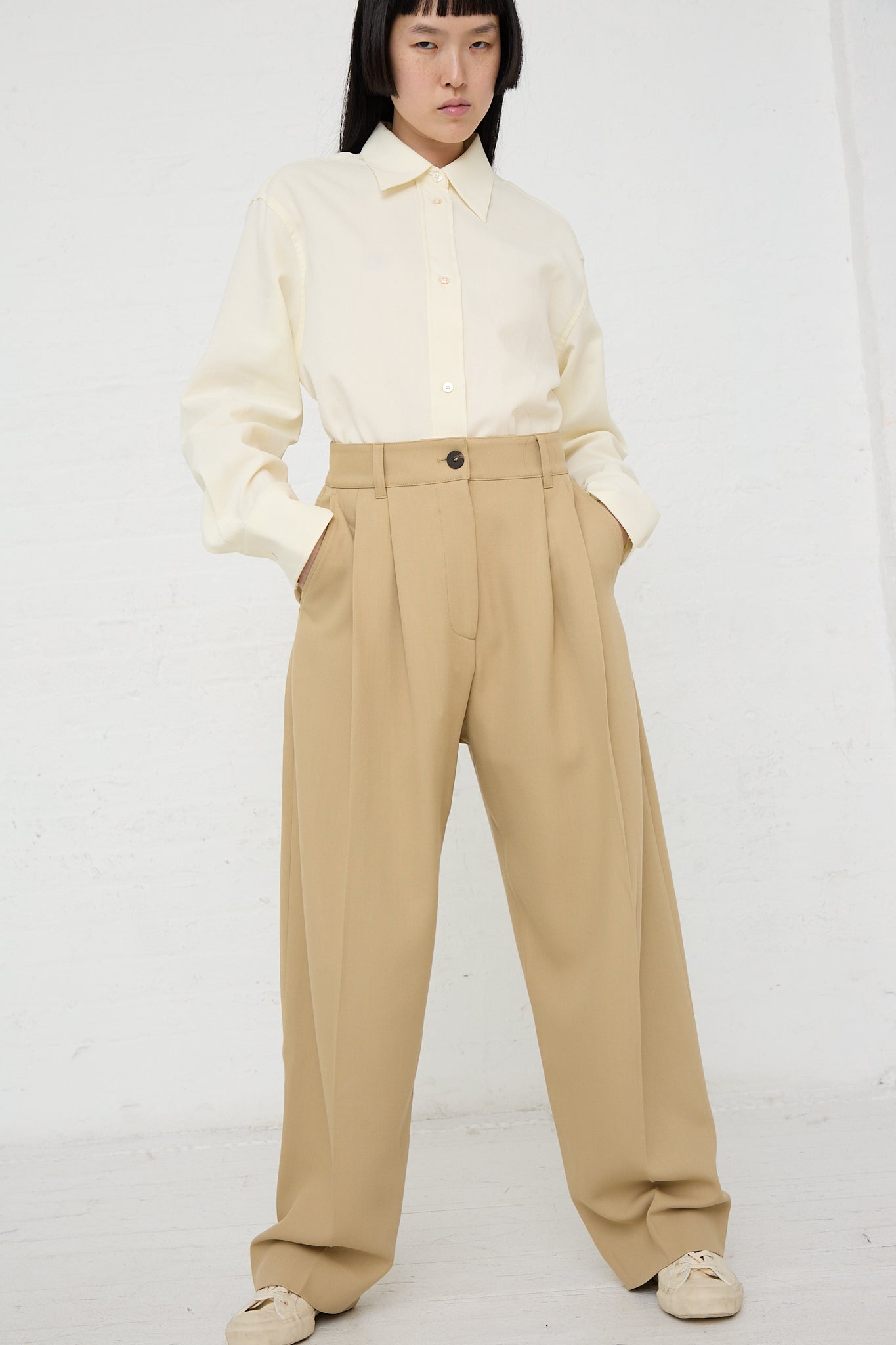 A model wearing Acuna Double Pleat Front Trouser in Sand and a cream colored button up blouse by Studio Nicholson. Full length and front view.