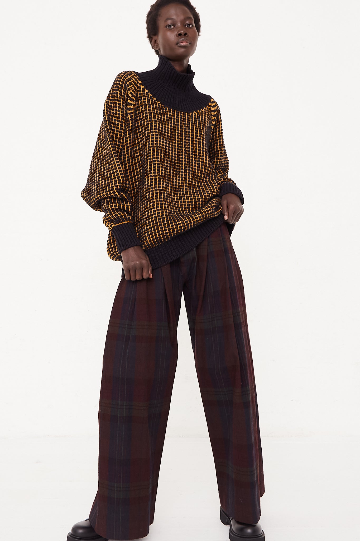 The model is wearing a black turtleneck sweater in Pitch Black Gold Lalin by Jan-Jan Van Essche and brown wide leg pants made of merino wool.