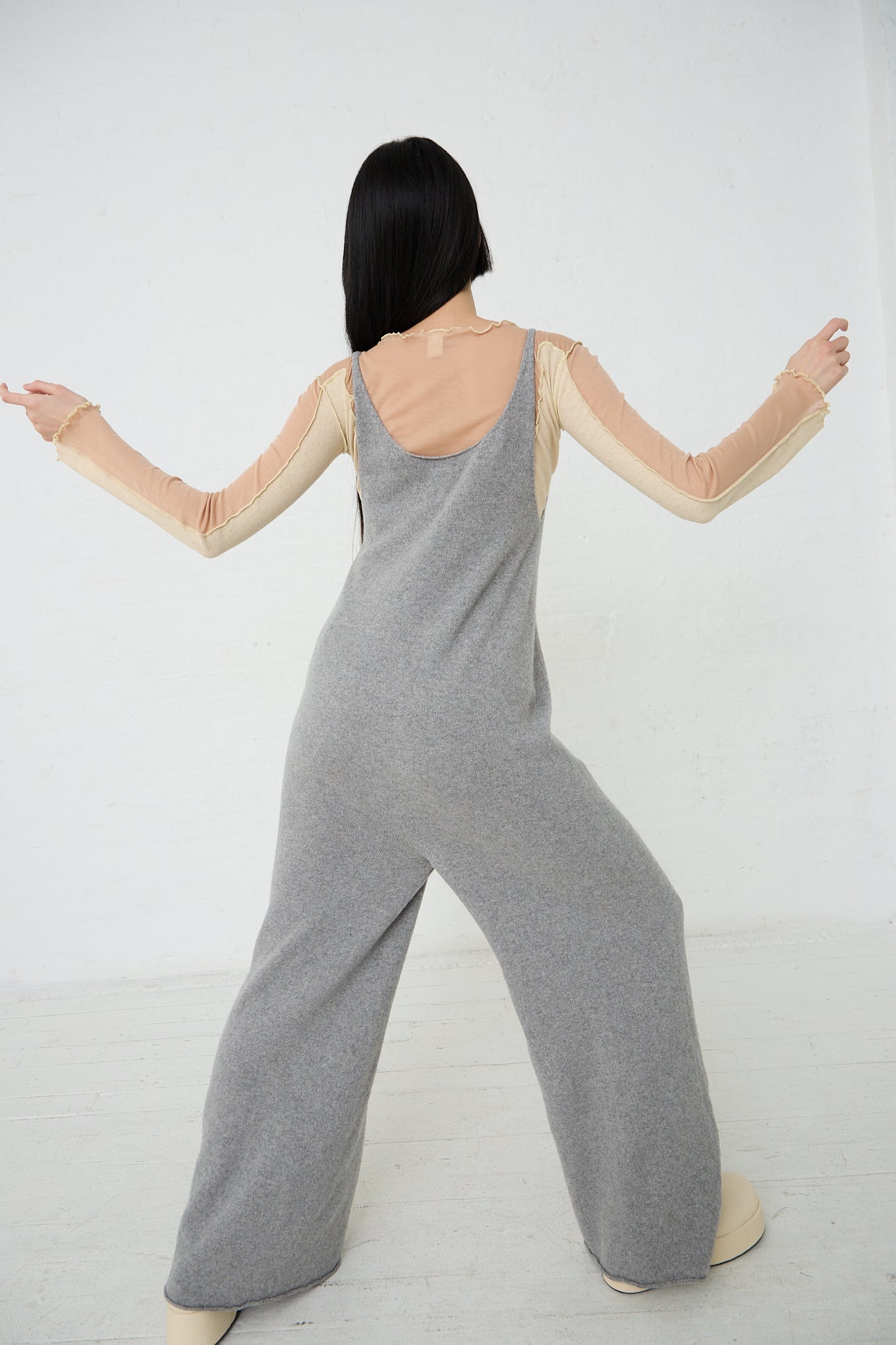 A woman in a Baserange Recycled Cashmere Rim Tank Jumpsuit in Grey Melange with her arms outstretched. Back view.