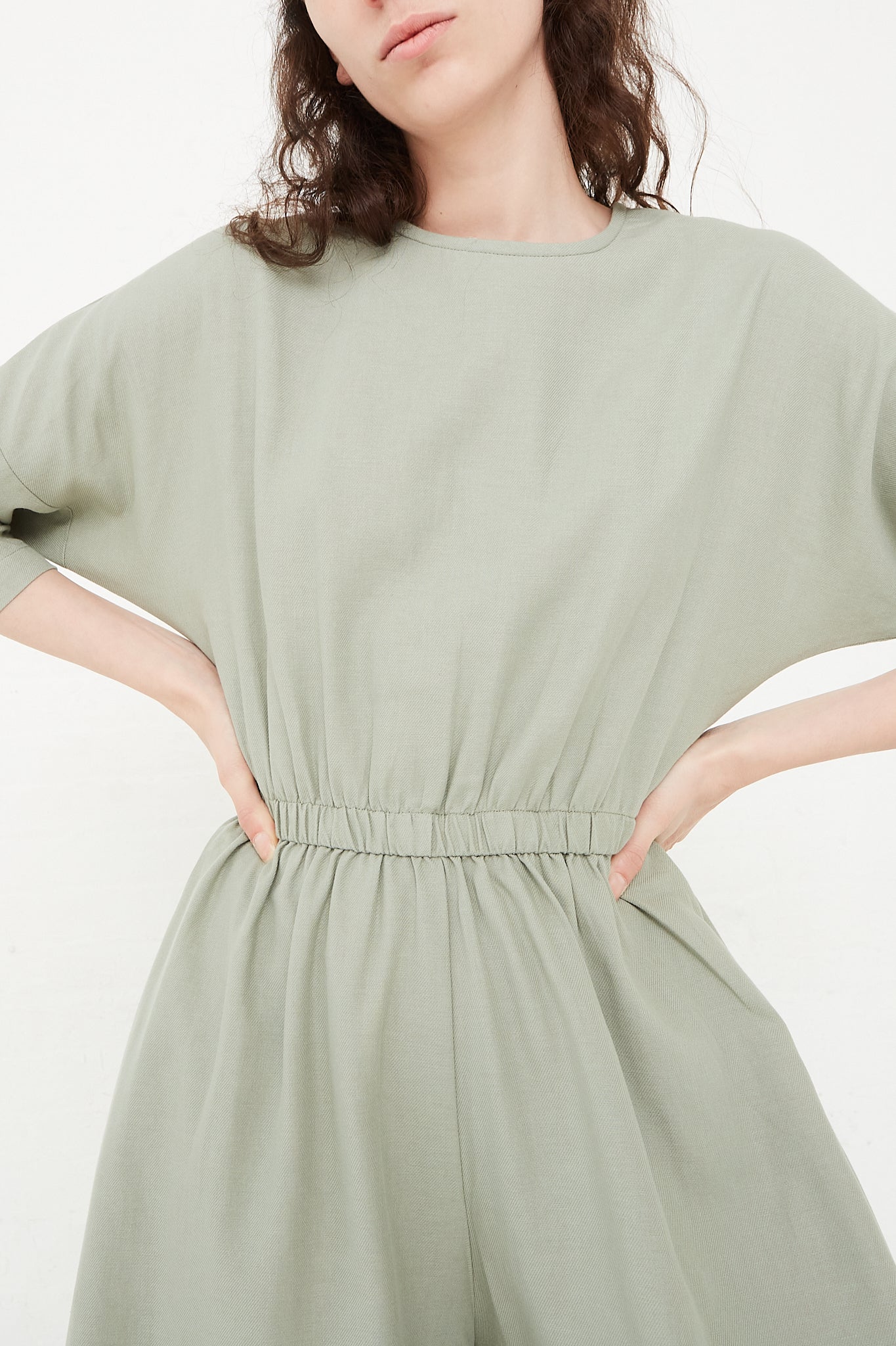 The model is wearing a sage green Cotton Twill Wide Culotte Jumpsuit in Agave by Black Crane with an elasticated waist and long sleeves.