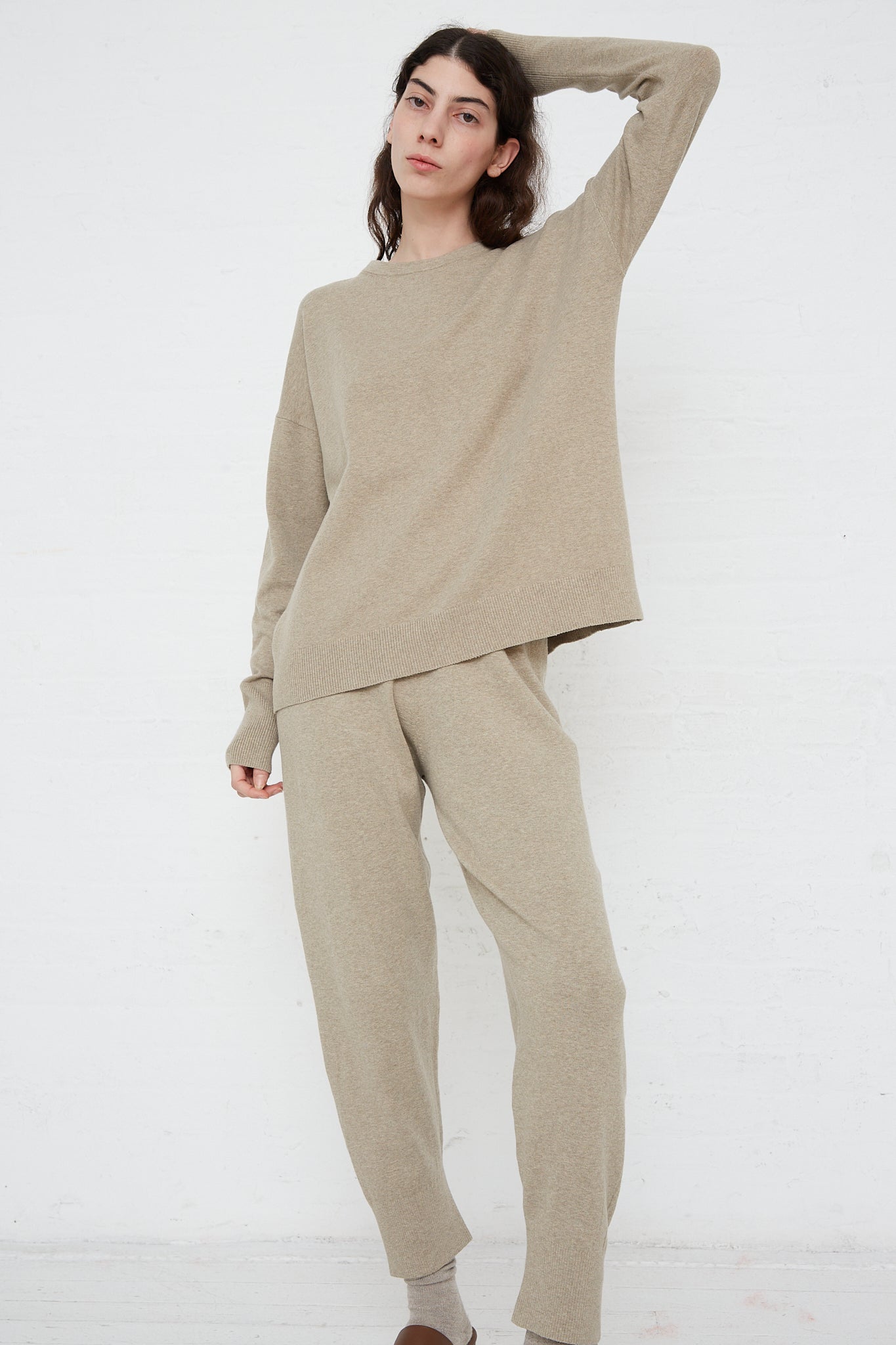 The model is wearing a beige sweater and relaxed fit joggers with an elasticated waist.Product: Lauren Manoogian's Base Pant in Stone Melange. Front view with model's left arm raised above head.