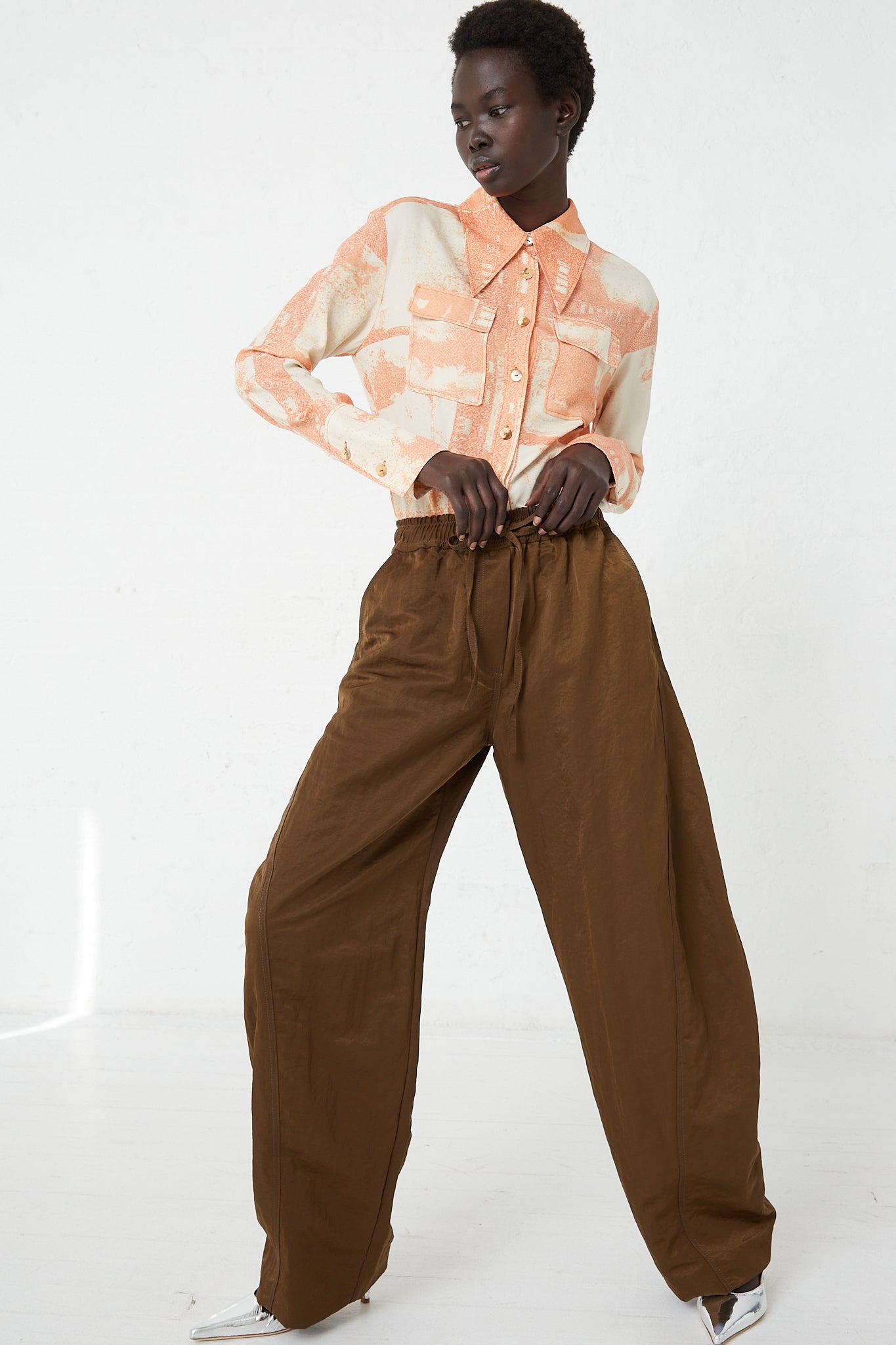 A model wearing Rejina Pyo's Nylon Una Trousers in Brown with contrast stitching and an orange shirt.