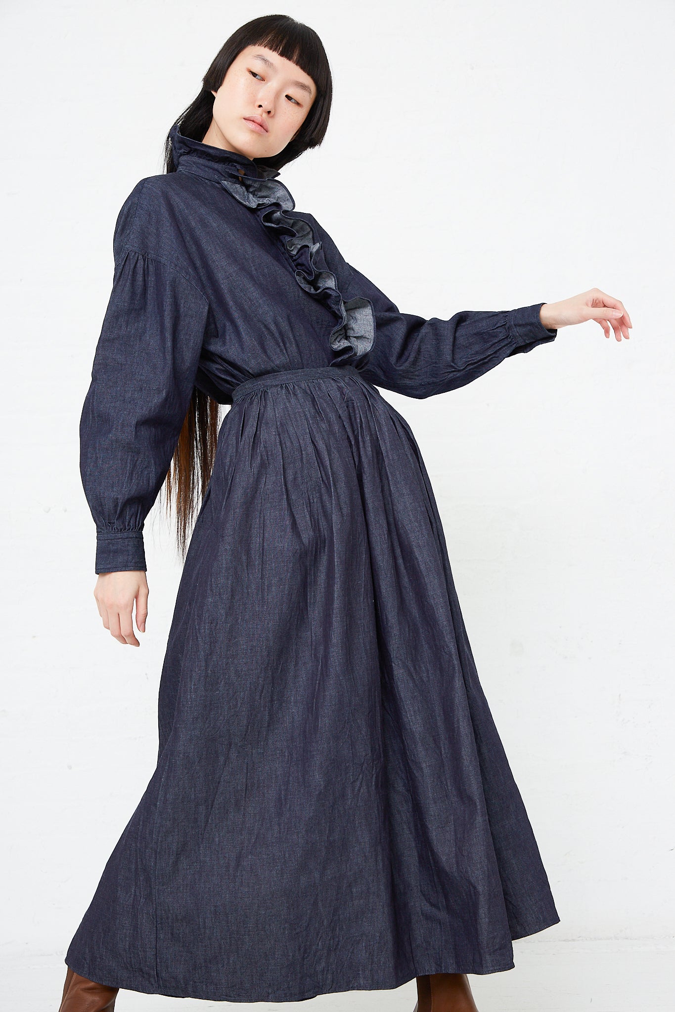 A woman wearing a Toujours Cotton Denim Cloth Pleated Maxi Skirt in Indigo with pleat gathers and ruffled sleeves.