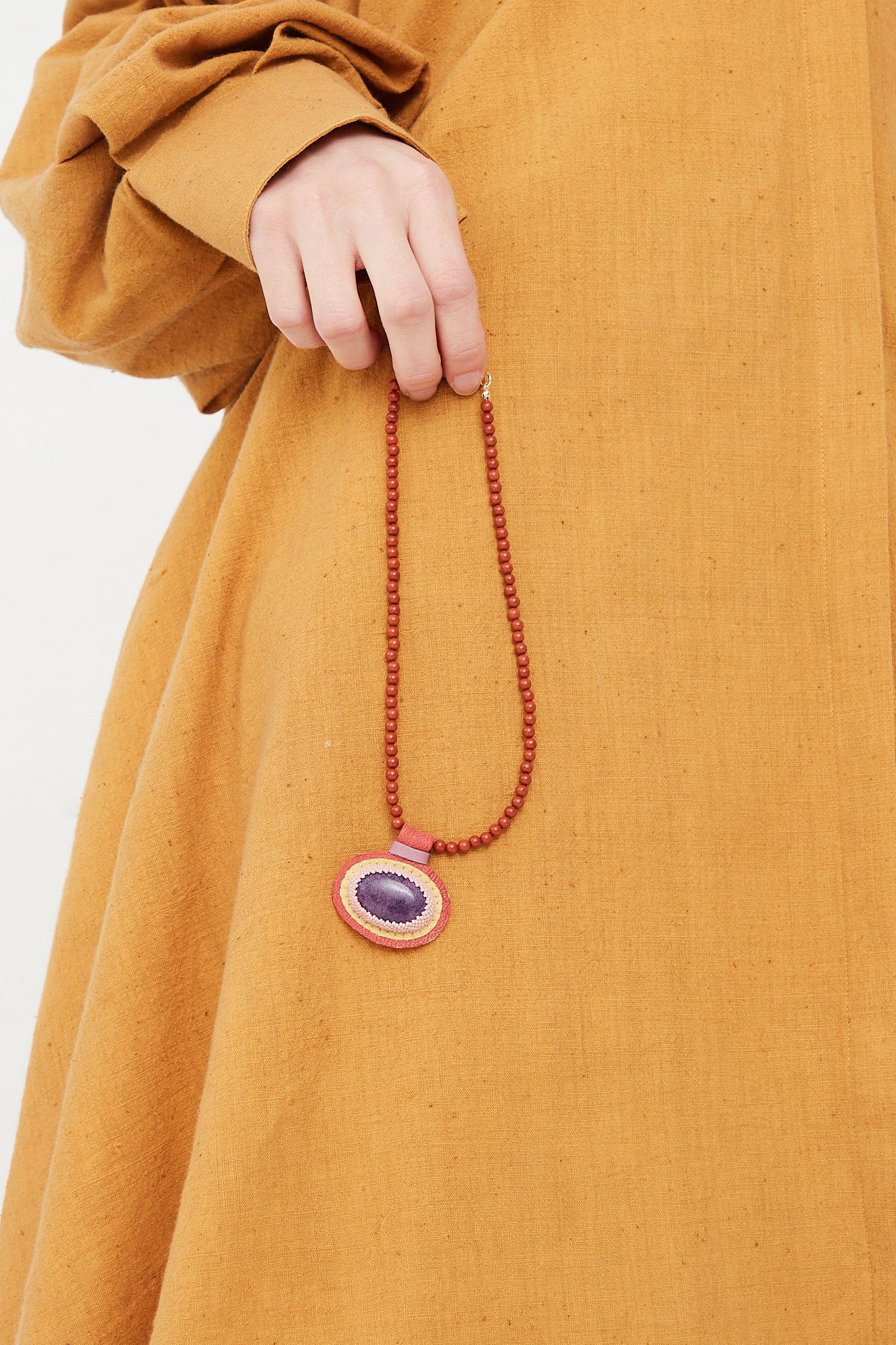 A woman wearing a yellow dress holding a Robin Mollicone Charm Necklace adorned with Red Jasper Beads and a Purpurite Charm.