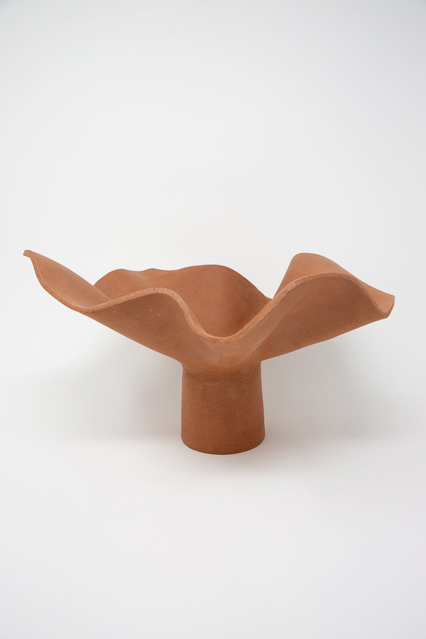 A hand-built clay sculpture, the Ruffle Pedestal in Terracotta by Lost Quarry is a ceramic flower-shaped bowl with a wavy rim, gracefully standing on a cylindrical pedestal base against a plain white background.