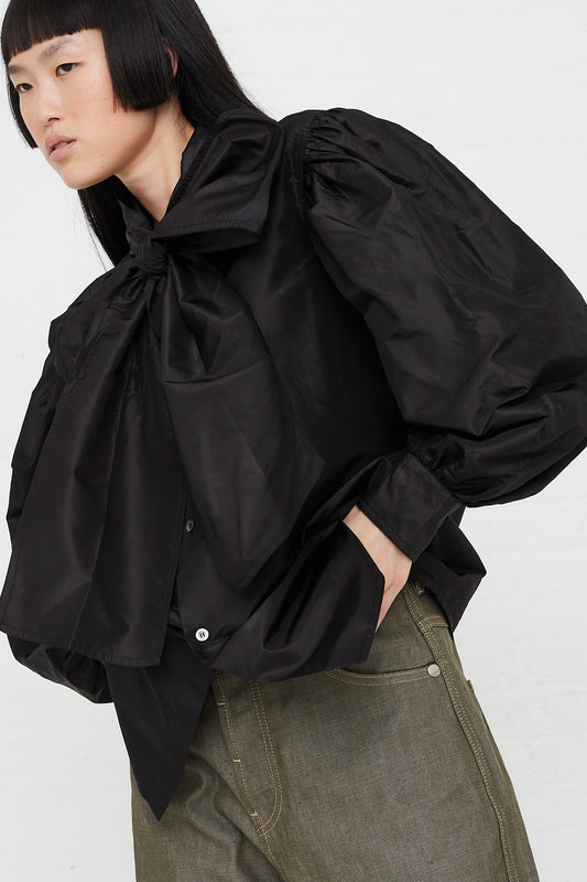 A woman wearing an oversized Sofie D'Hoore Silk Taffeta Burnette Shirt in Woven Black with a bow. Side profile.