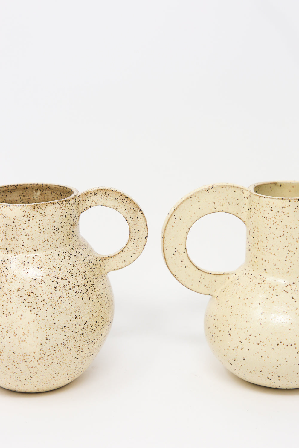 Two "Arches in Sun Drop" jugs by Lost Quarry, featuring speckled off-white ceramic with round bodies and large handles crafted from speckled clay, stand side by side against a plain white background. Perfect as medium vessels for floral arrangements.