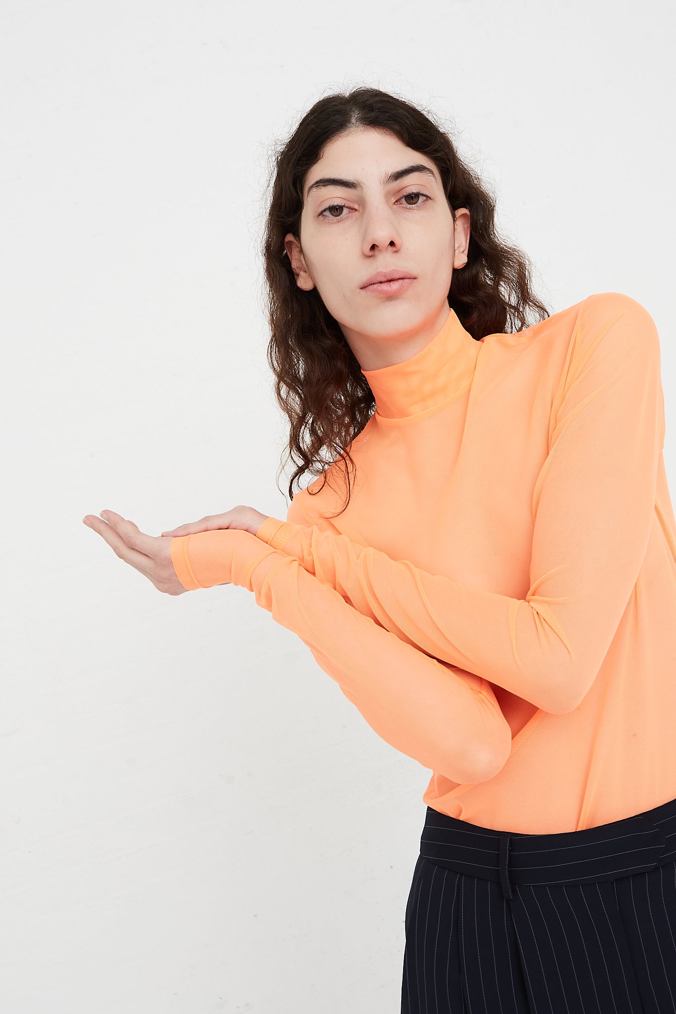 A model wearing a Nomia Long Sleeve Mesh Mockneck in Fluoro Orange top and black pants, designed by the NOMIA brand, at Oroboro Store.