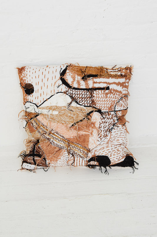 A Complex Embroidery Cave Cushion by Luna Del Pinal, handmade in brown and black with intricate hand embroidery, is placed on a white wall.