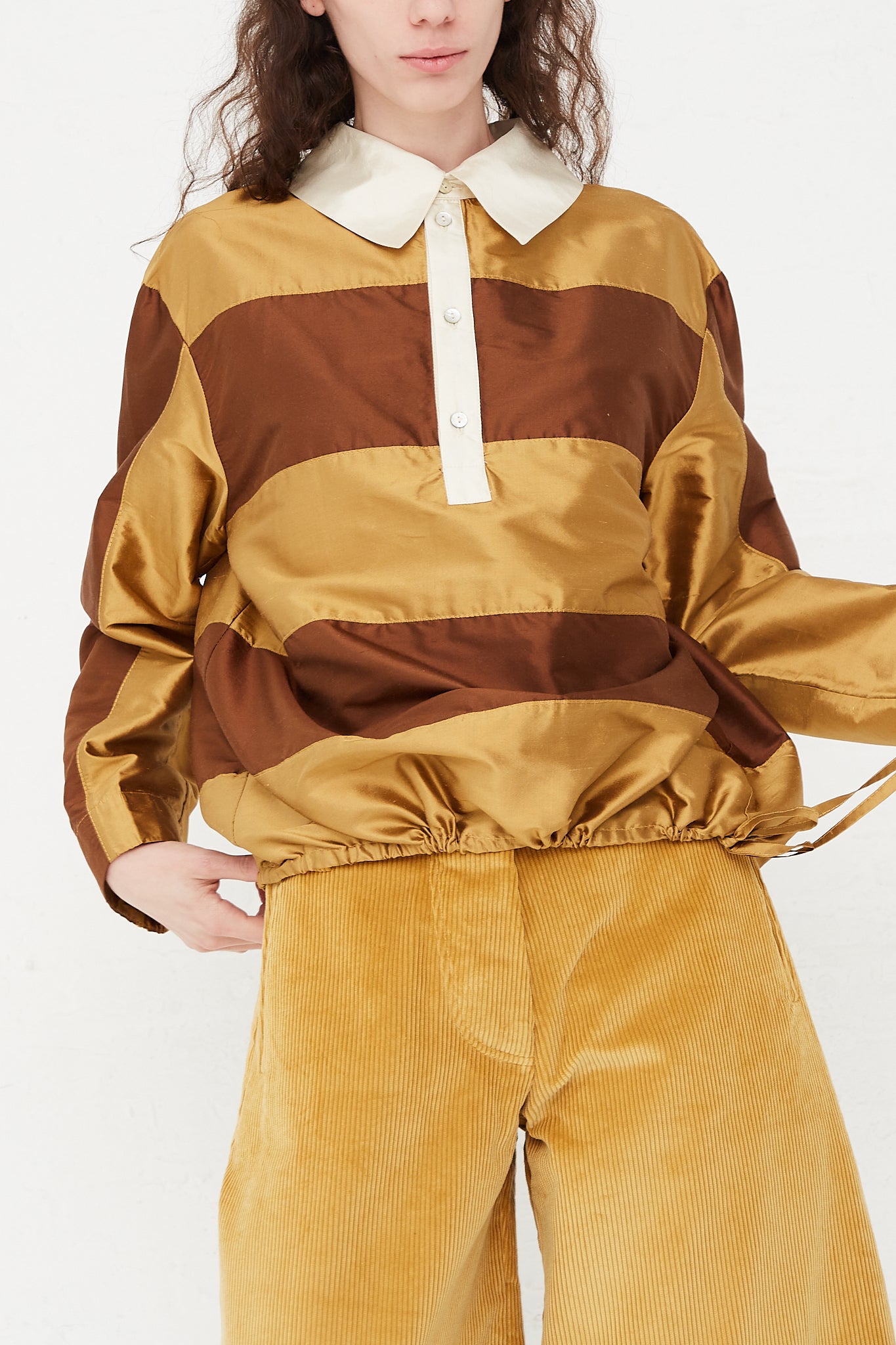 A model wearing a striped rugby shirt in a shiny silk doupion. Features a contrast fold-over collar and button placket, with an adjustable drawstring at hem. Front view and up close. Highlighting drawstring detail. Designed by Cawley - Oroboro Store 