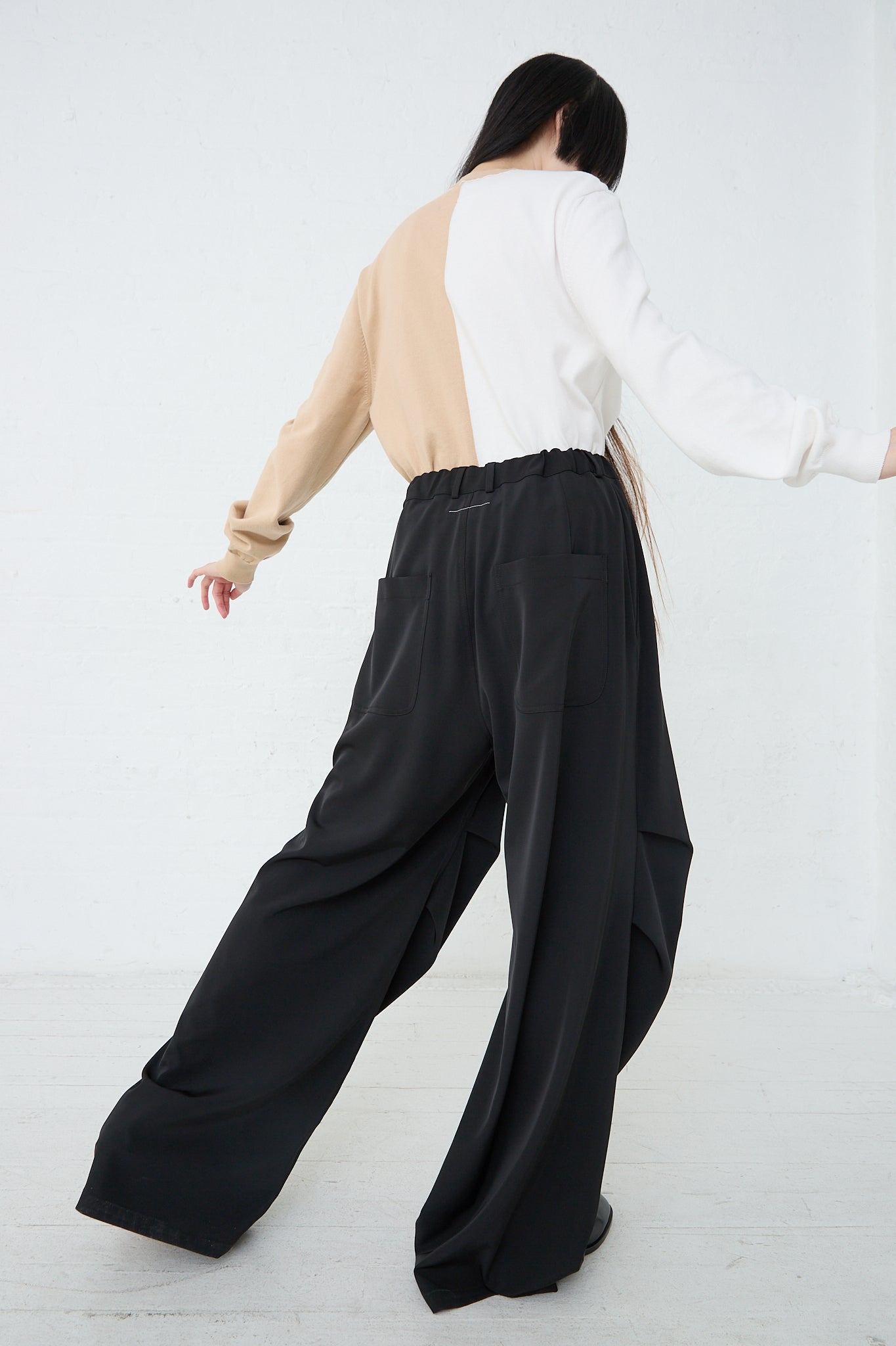 A woman is standing in a white room wearing MM6 black twill stretch pants with an oversized silhouette. Back view and full length.