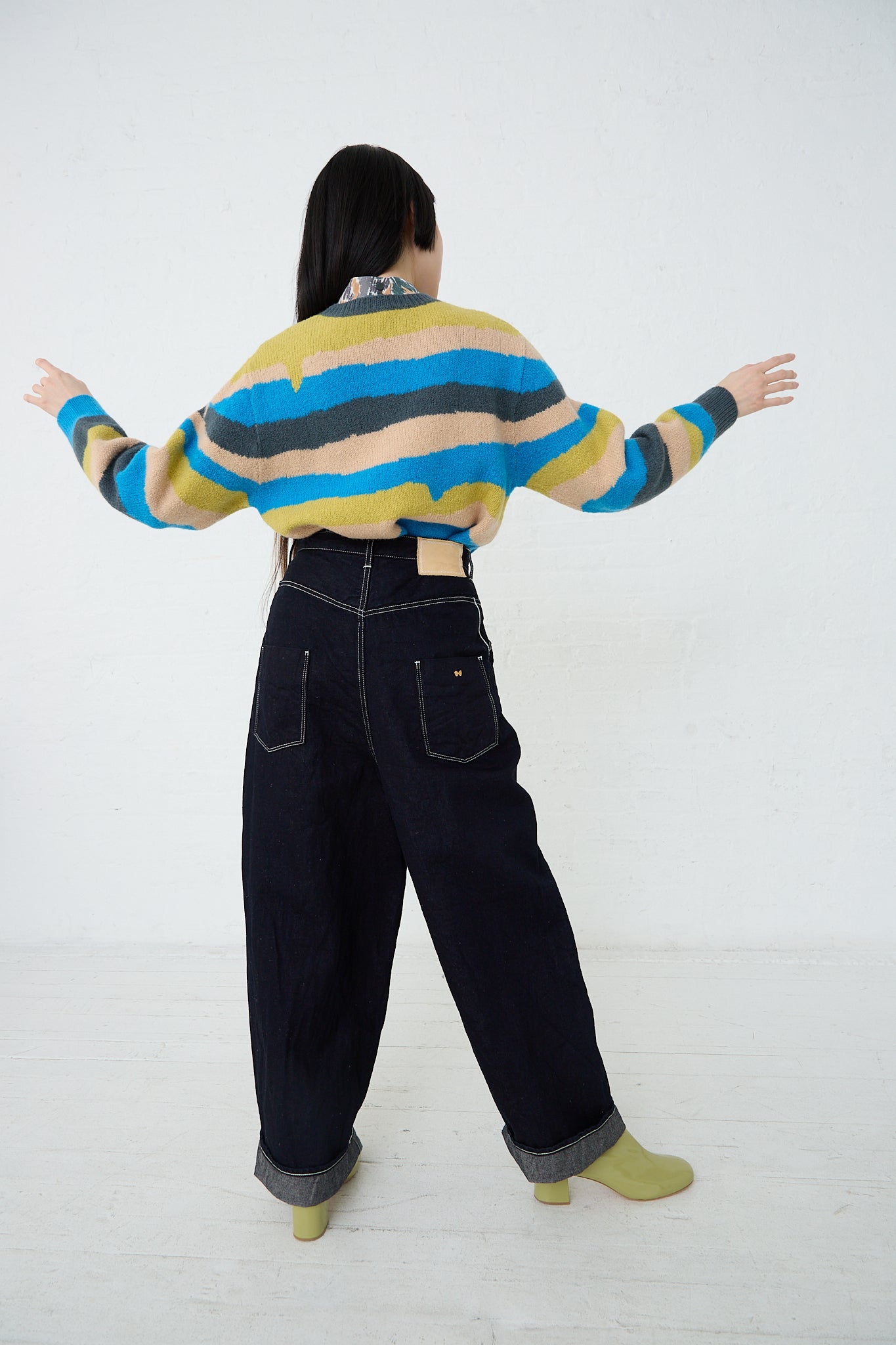 A woman is standing in a white room with her arms outstretched, wearing the Mina Perhonen Always Balloon Wide Pant in Indigo. Back view and full length.
