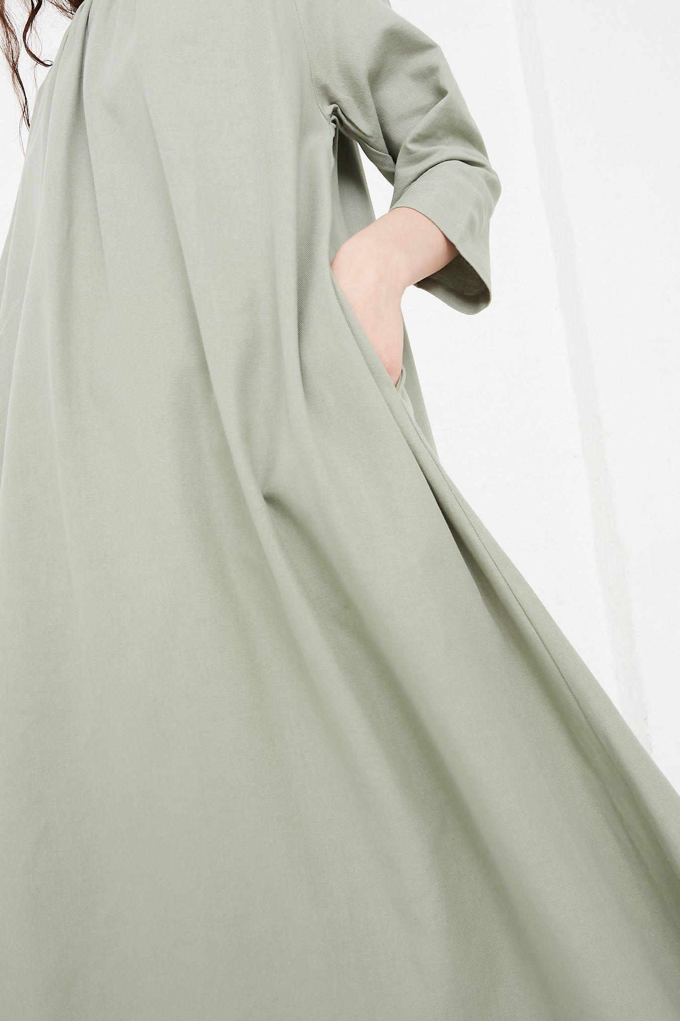 The model is wearing a Cotton Twill Shirred Neck Dress in Agave by Black Crane. Side view. Model's hand is in pocket.
