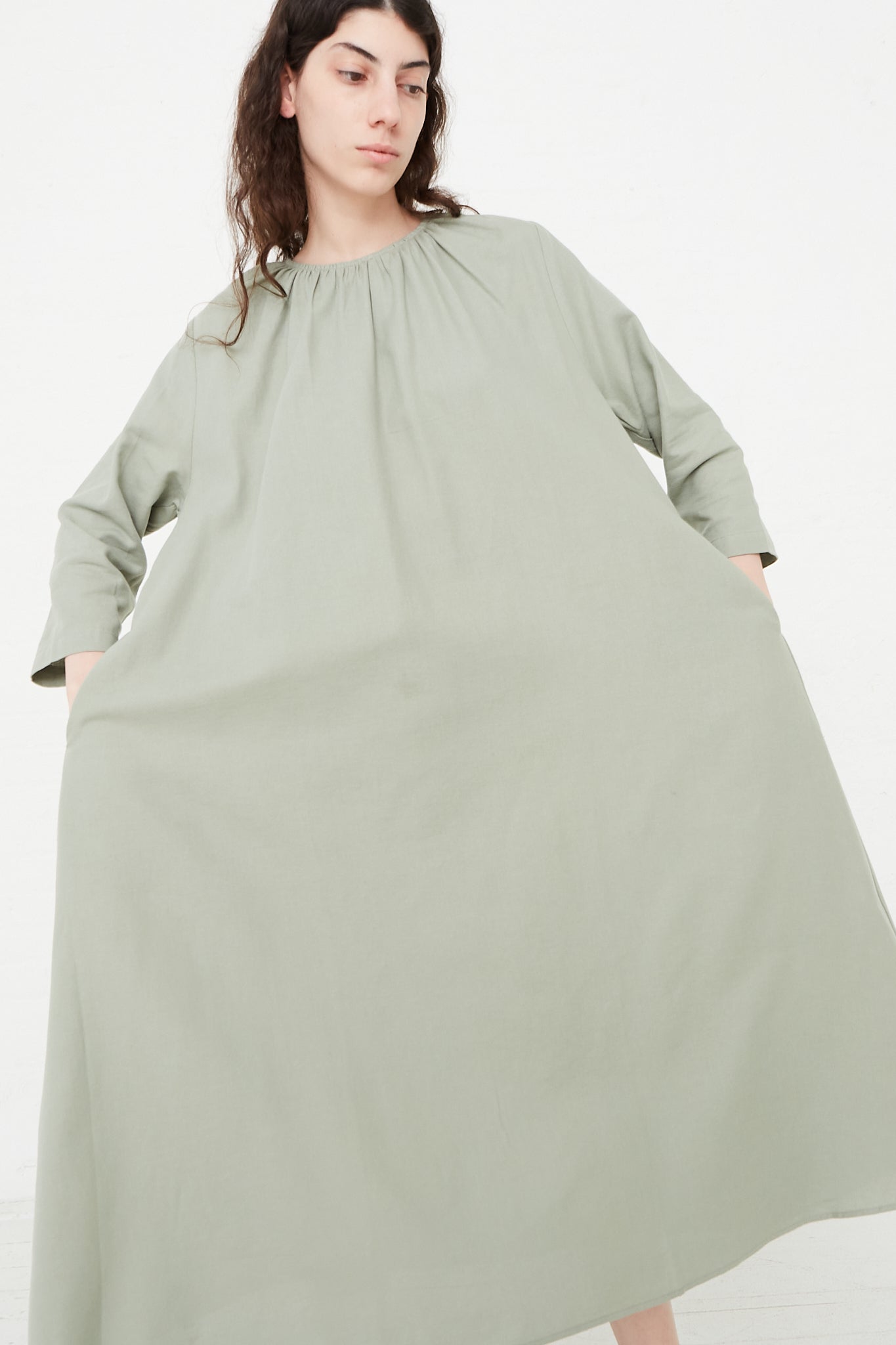 The model is wearing a sage green Cotton Twill Shirred Neck Dress in Agave by Black Crane. Front view. Model's hands are in pockets.