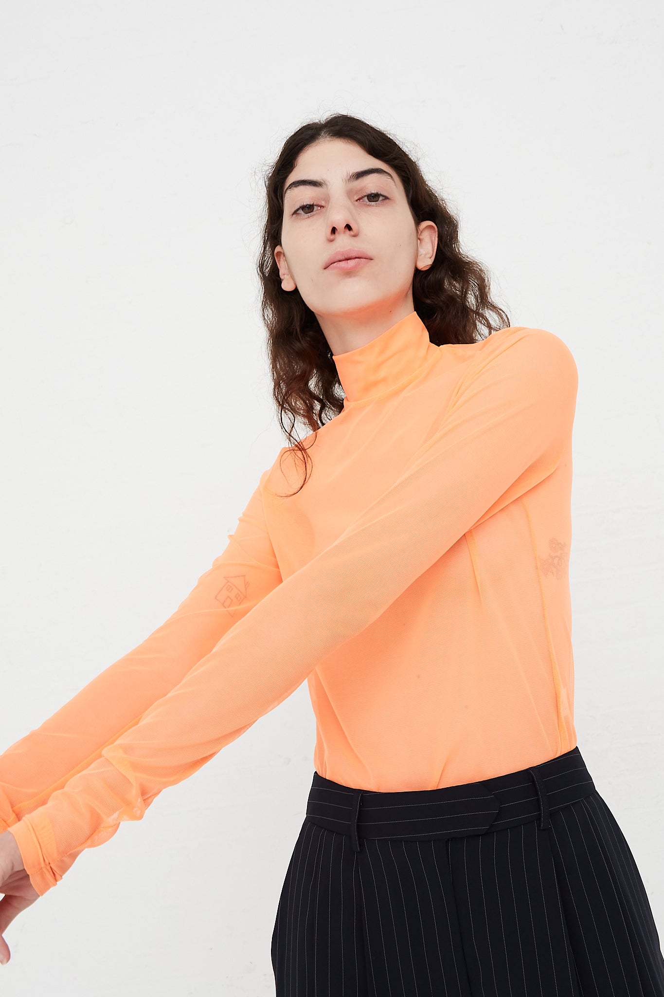A model wearing a Long Sleeve Mesh Mockneck in Fluoro Orange top and black pants designed by Nomia brand.