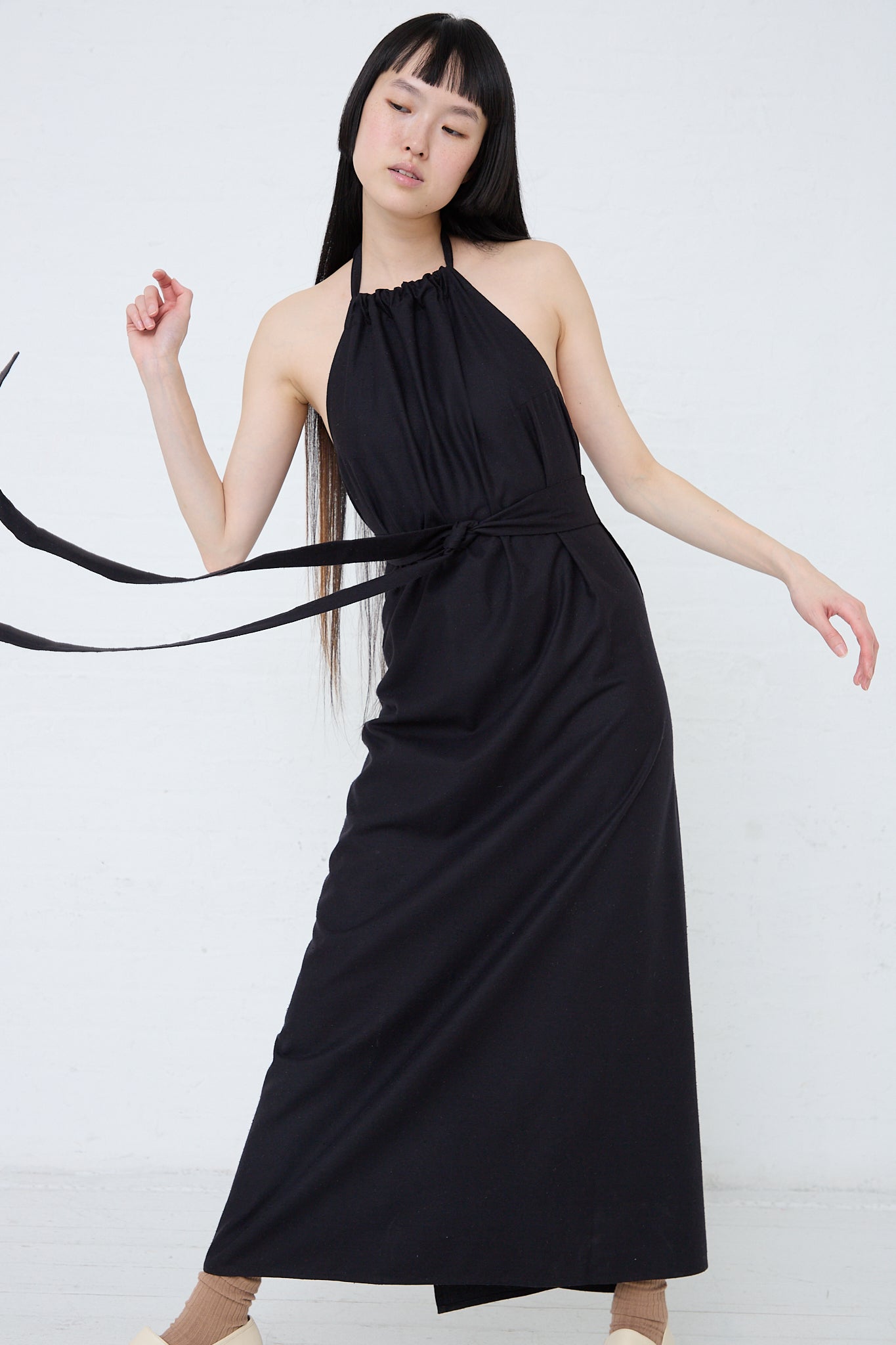 A woman in a Baserange Wild Silk Trope Apron Dress in Black is posing. Front view and full length.