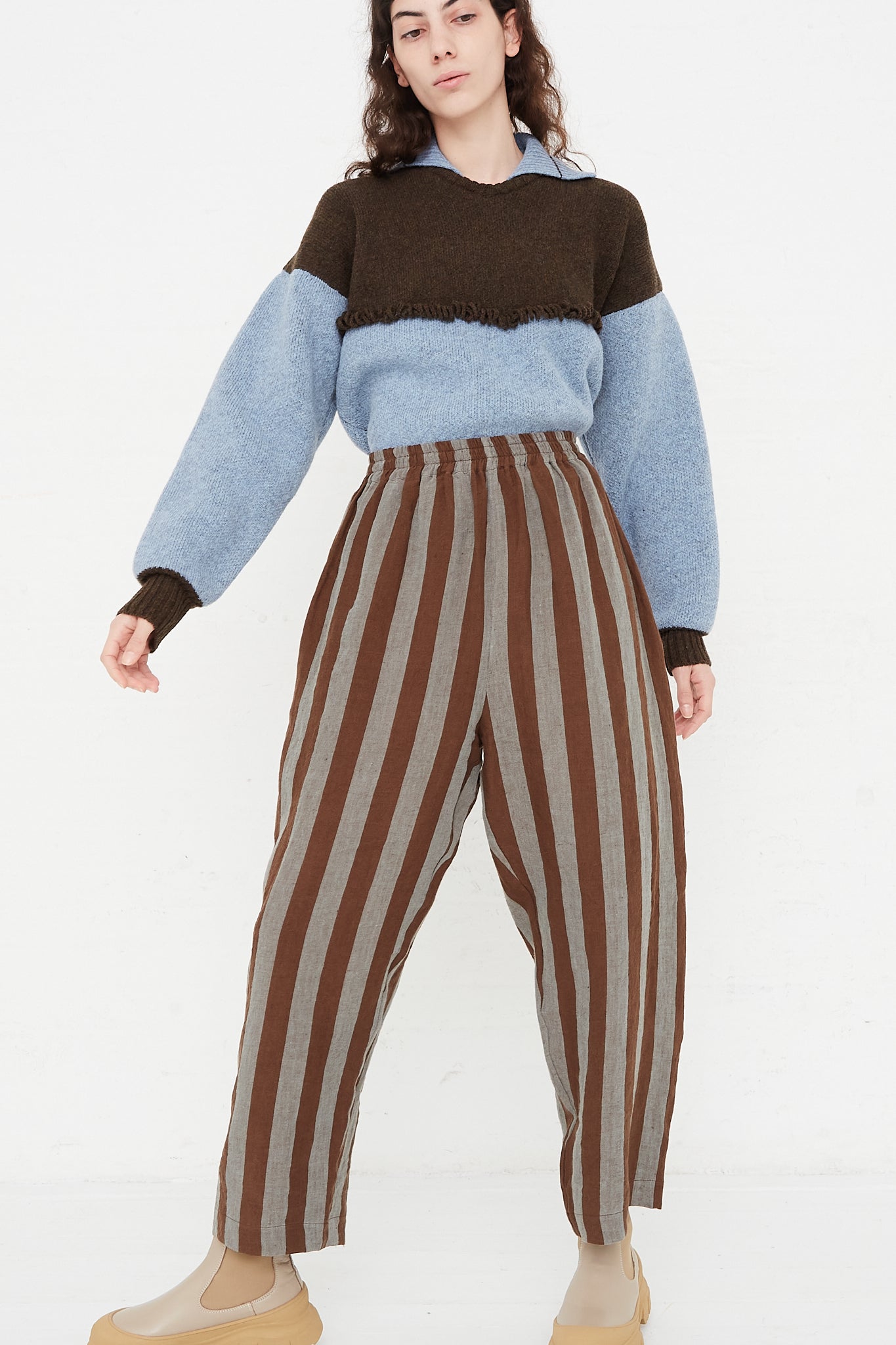 A model wearing a high waist trouser in a striped Irish linen. Brown and blue stripe. Features an elasticated waist and a wide straight leg. Front and full view of model highlight full pant, sweater and boots. Designed by Cawley - Oroboro Store