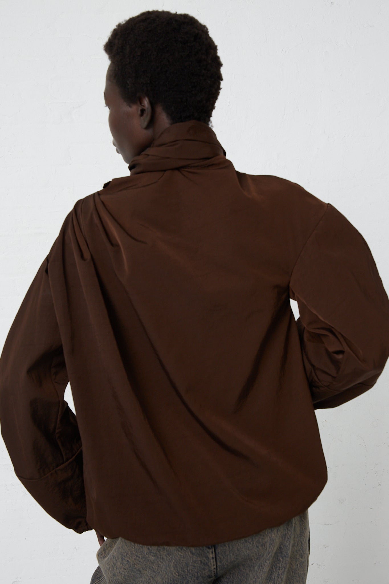 A woman wearing a Veronique Leroy Zipped on Sleeve Rain Blouse in Choco with a removable hood. Back view.