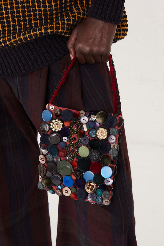 A handmade bag with mixed buttons on vintage fabric. Features a snap closure and beaded strap | Intensity - Oroboro Store | Up close of model holding bag against leg. Showing strap and hand.