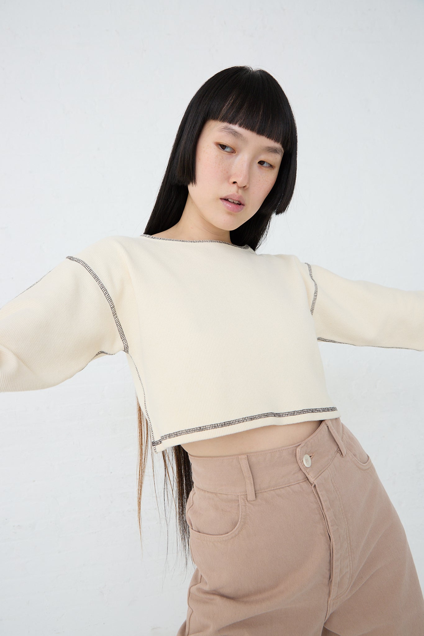 A woman wearing a Cotton Hemp Rib Garble Top in Undyed made by Baserange and tan pants. Front view. Model's arms are raised.