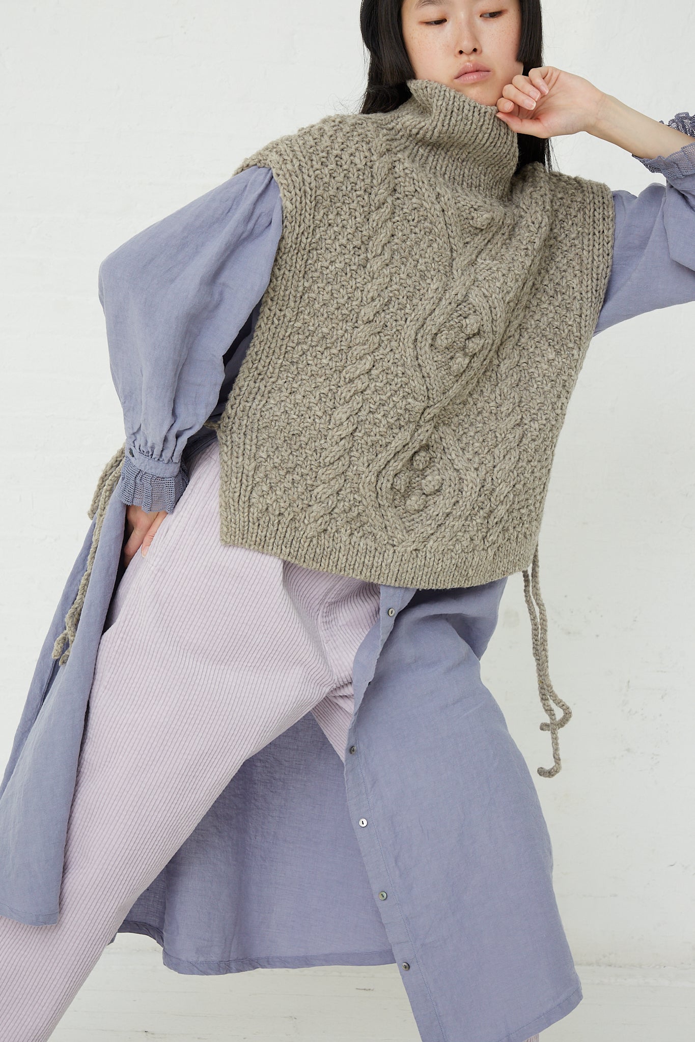 A woman wearing a nest Robe Peruvian Wool Hand Knitted Cable Stitch Vest in Beige and purple pants.