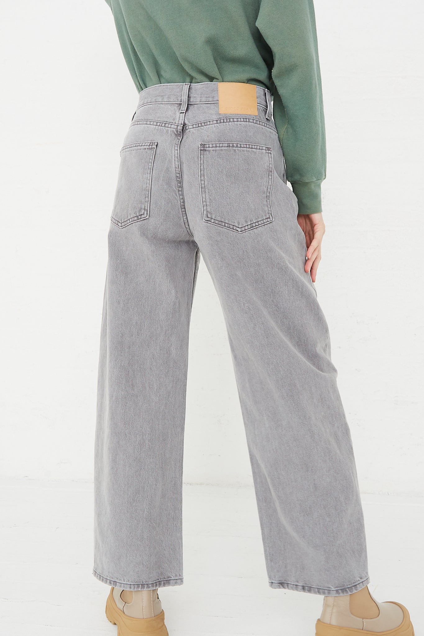A back view of a model wearing the "Leroy Mid Relaxed Bow Jean in Grey Stone" by B Sides and a green sweater.