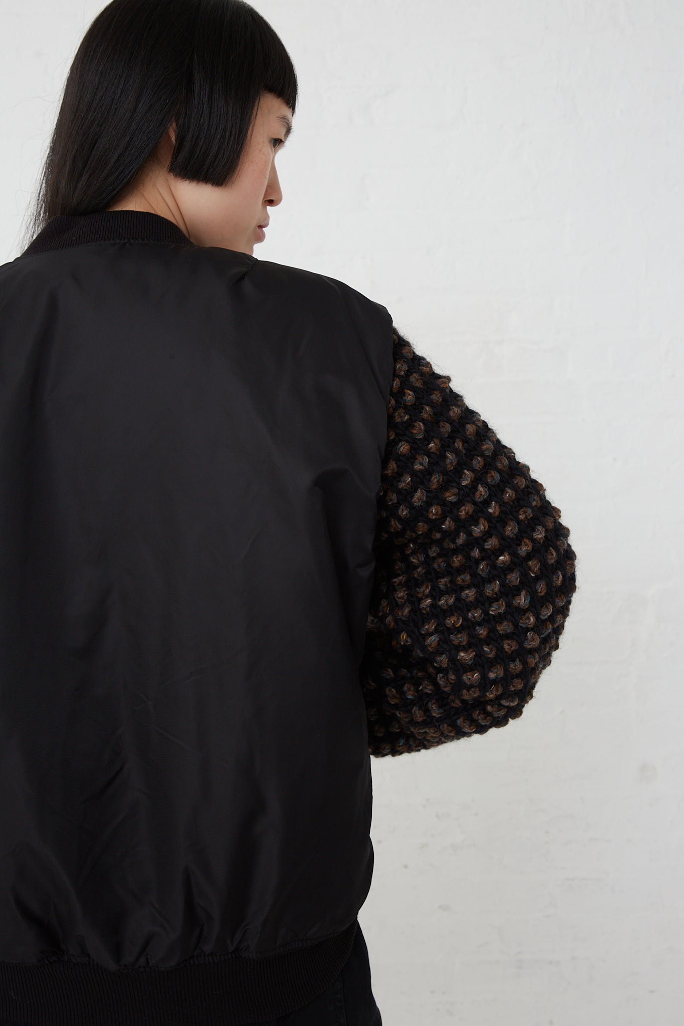The woman dons an oversized black Sleeve Bomber No. 70 made from a nylon and wool blend fabric. Back view.