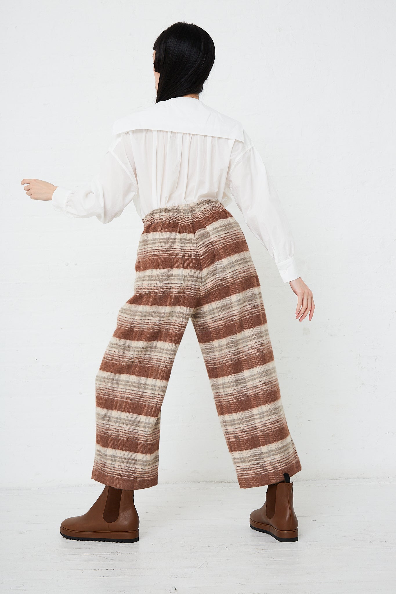 A woman in Silk Plaid Mosser Cloth Pajama Pants in Cocoa Brown by Toujours is standing in front of a white wall. Back view.