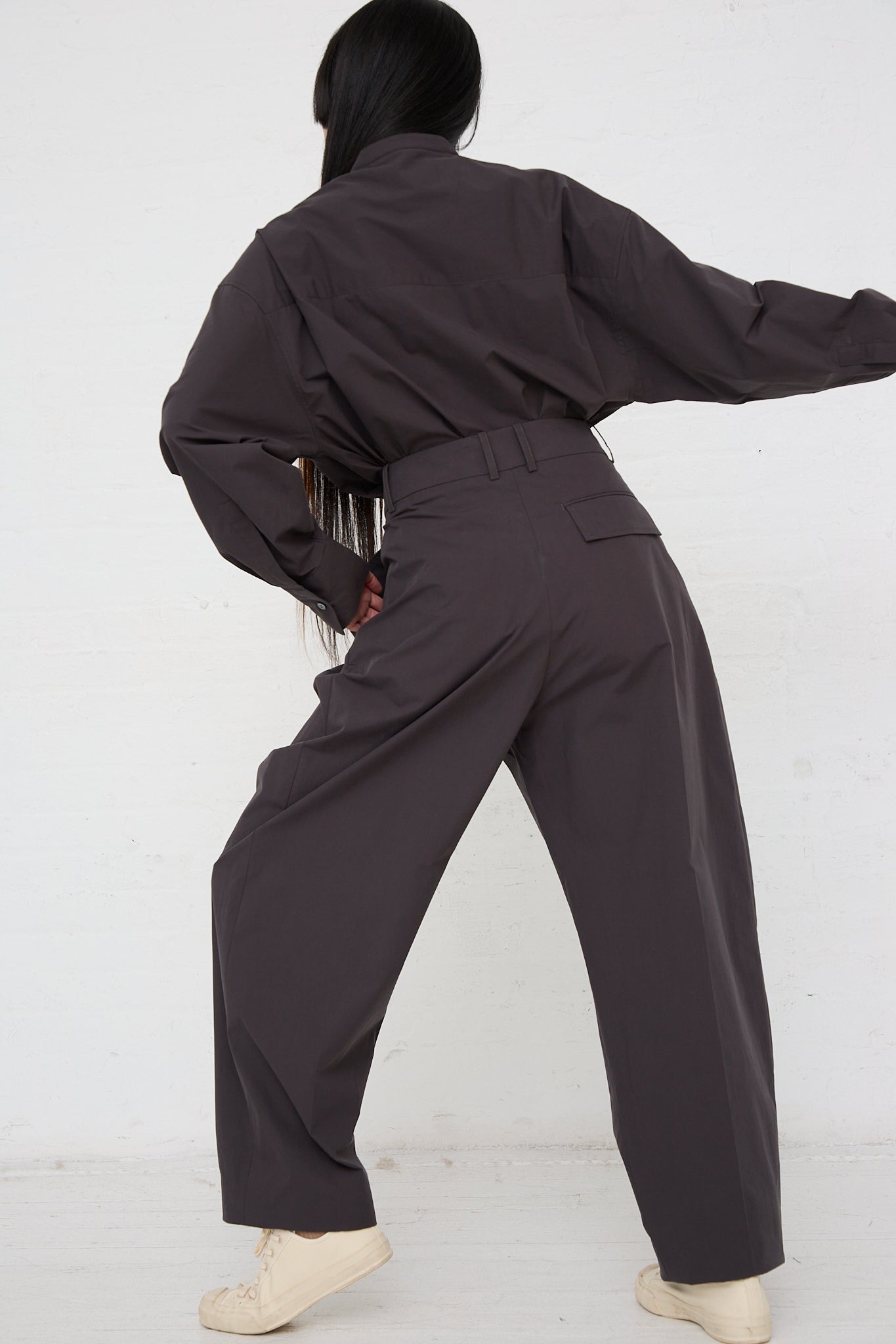 A woman in an Acuna Double Pleat Front Trouser in Asphalt by Studio Nicholson is standing in front of a white wall. Back view. Model's right arm is raised.
