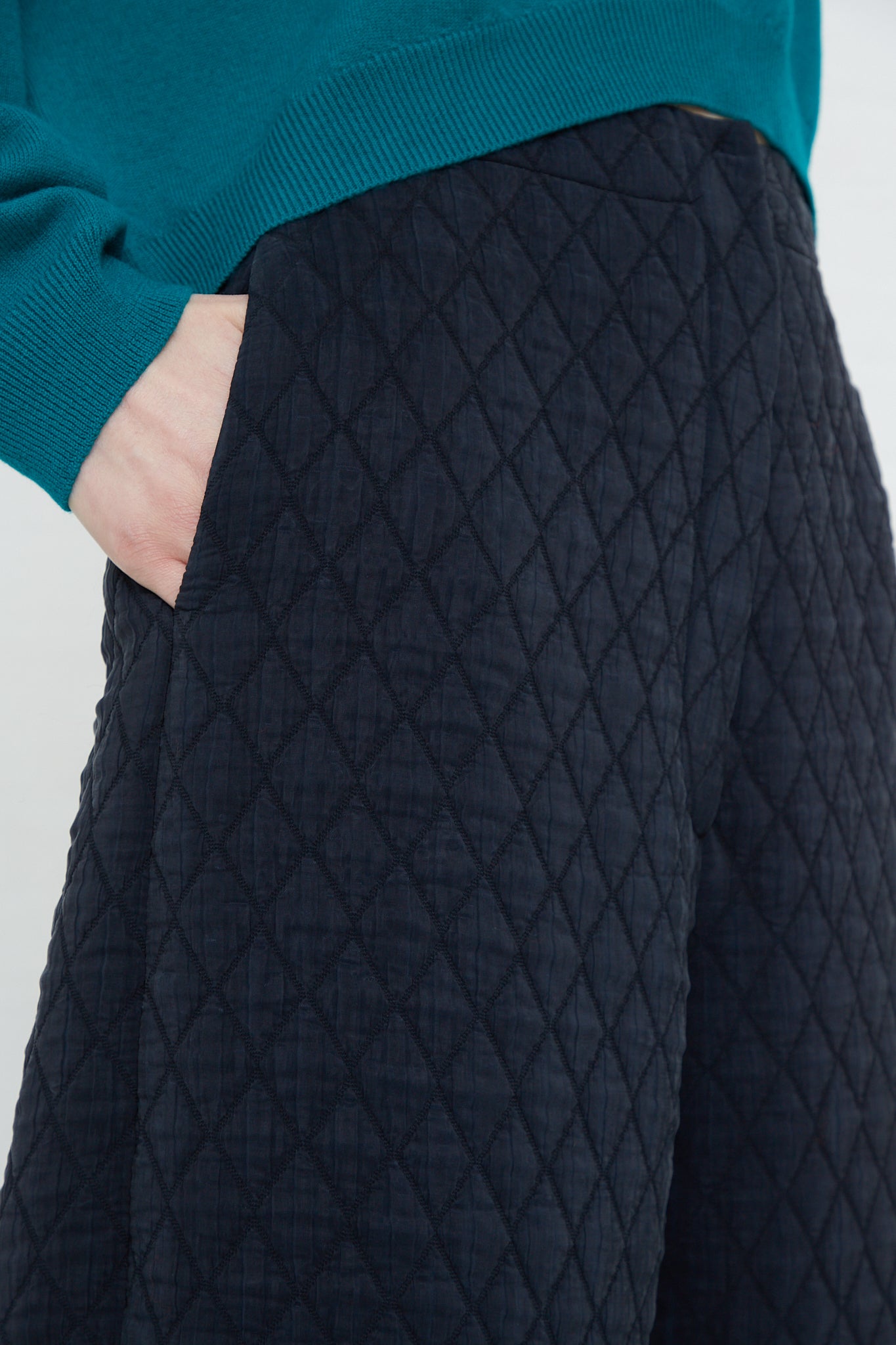 A woman wearing Cordera high-rise black Quilted Curved Pants with an elasticated waist and a teal sweater made from recycled polyester.