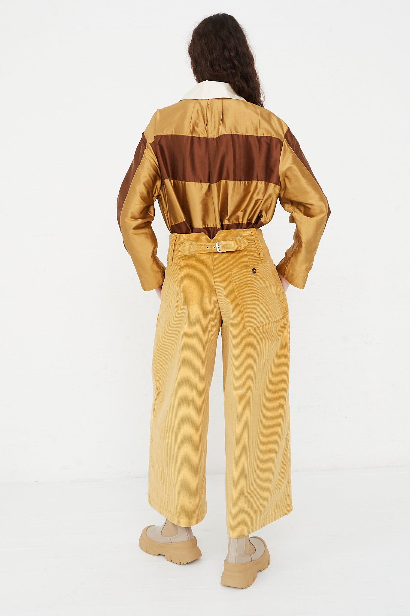 A model wearing a high waist trouser in a British cotton cord. Features a wide waistband, pleat detail and side welt pockets. Back view and full length. Hands in pockets. Designed by Cawley - Oroboro Store