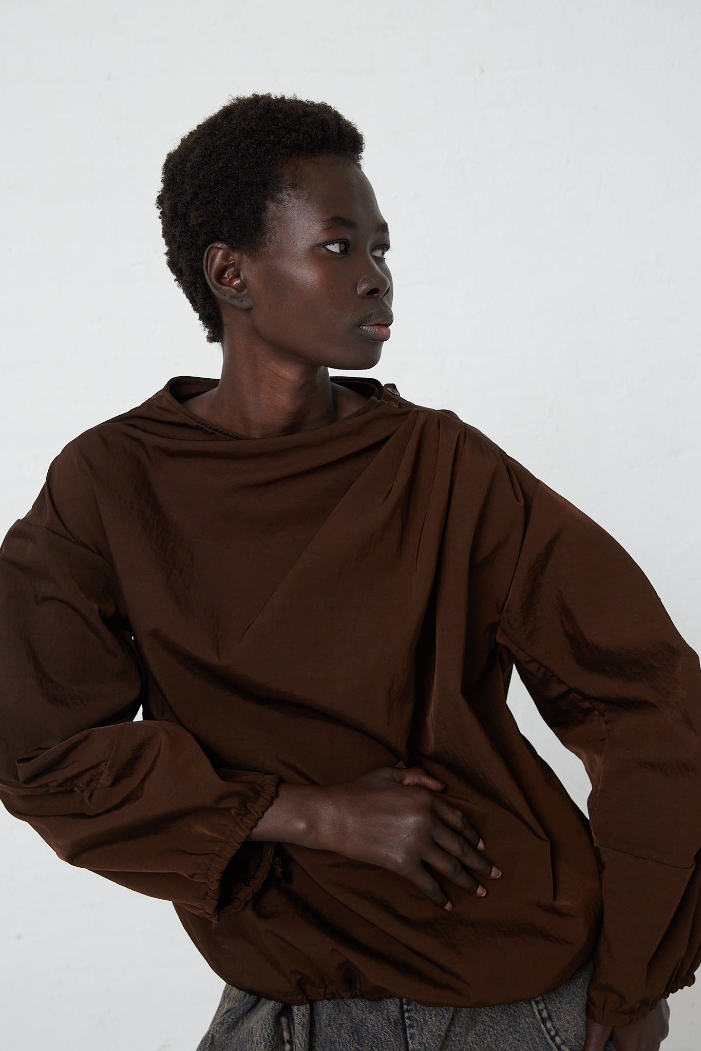 A woman is wearing the Veronique Leroy Zipped on Sleeve Rain Blouse in Choco, which has a removable hood and is made of taffeta nylon material.